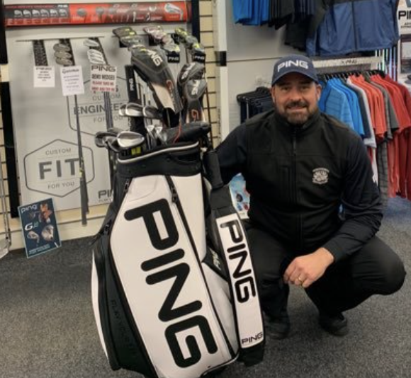 Man kneeling next to a PING golf bag with golf clubs inside at a sporting goods store.