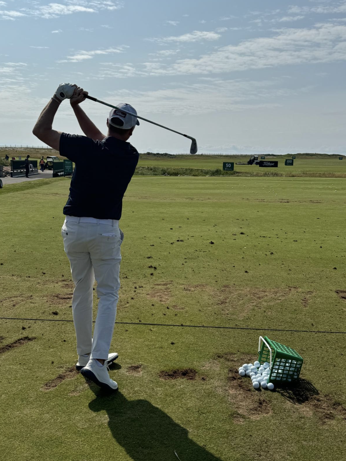 A man preparing to hit a golf ball on a driving range with a golf club, with a basket of golf balls next to him and other golfers practicing in the background.
