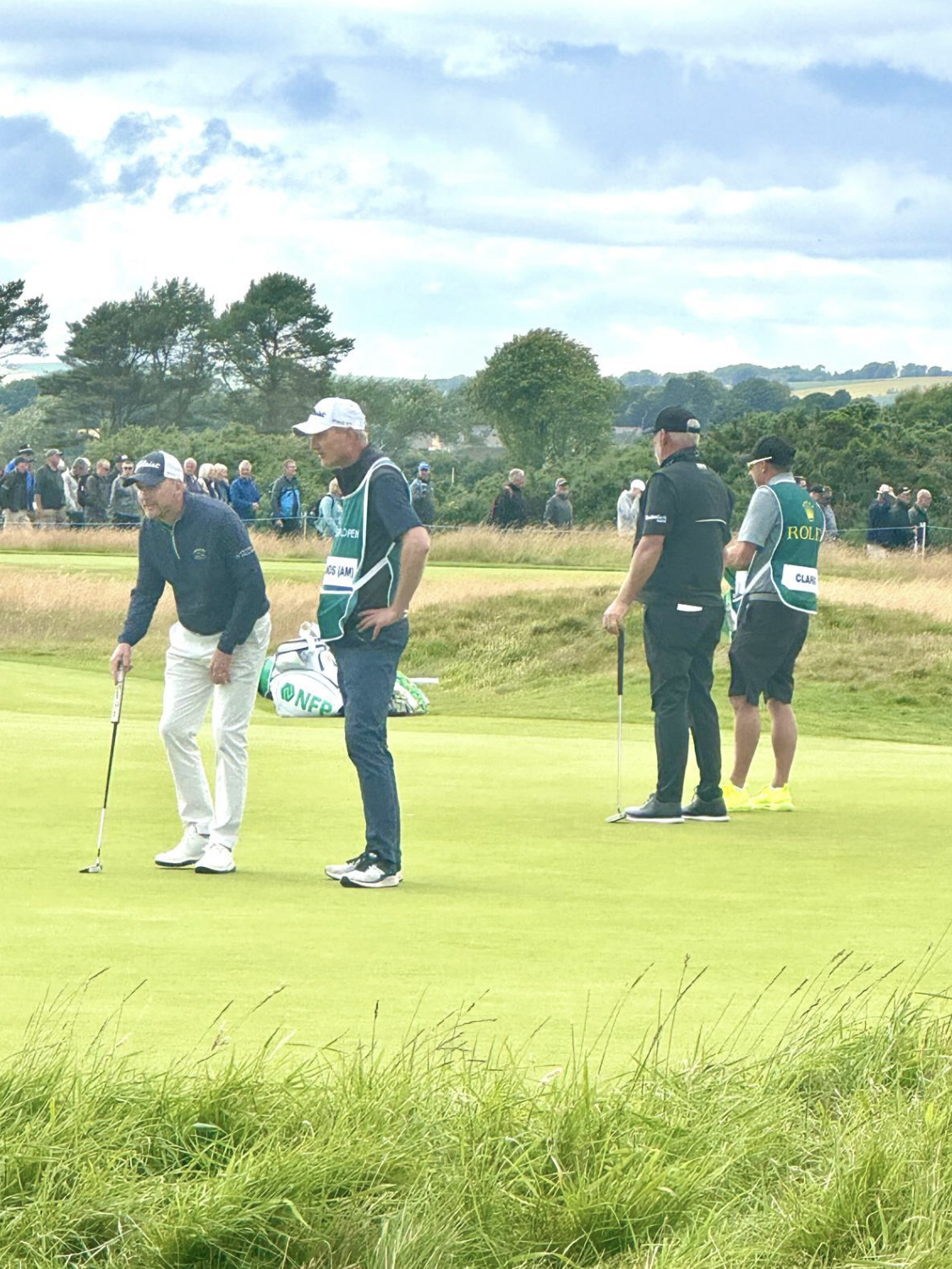 Golf players and officials on the green during a golf tournament, with spectators in the background under cloudy skies.