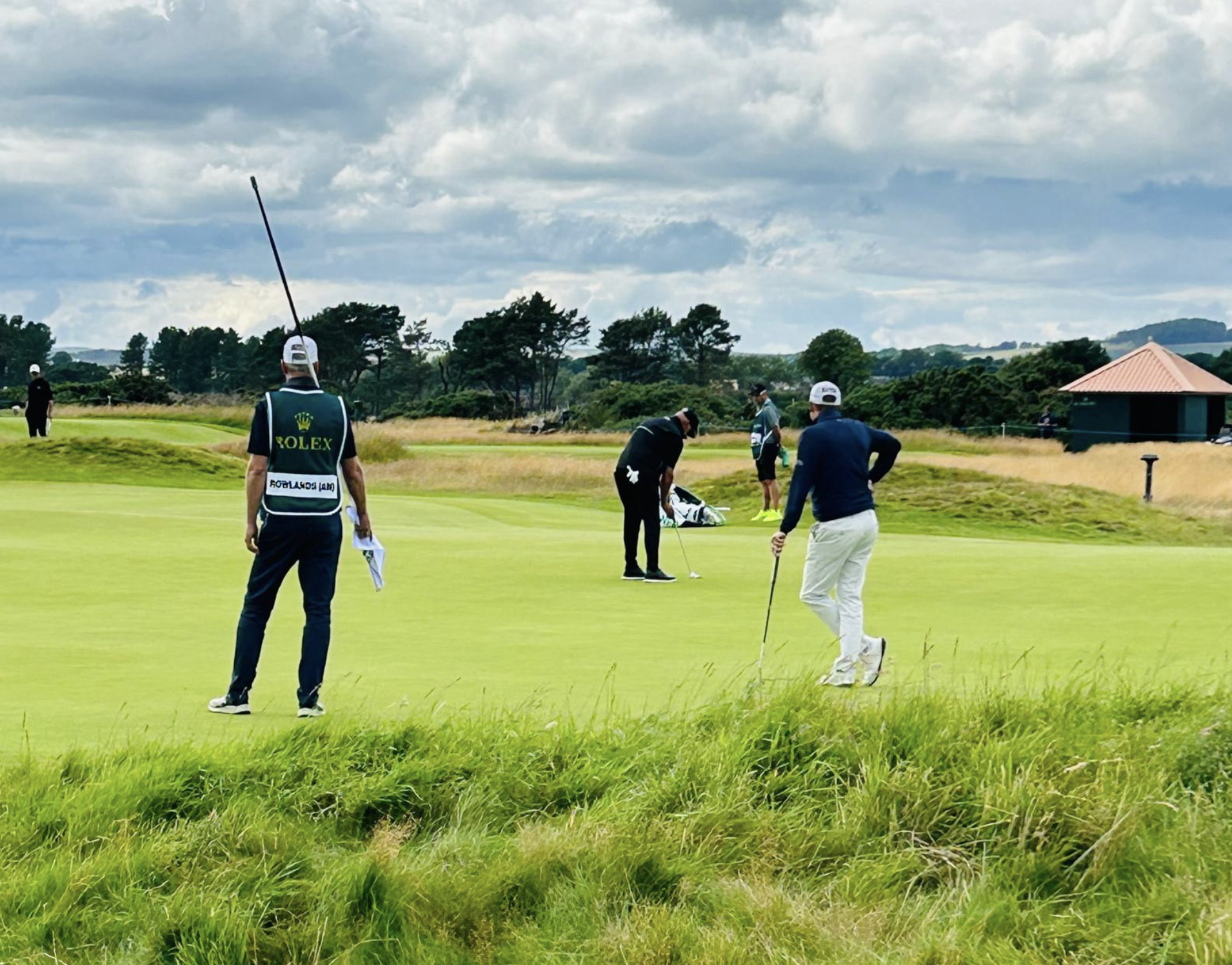 Golfers on a course with green grass, trees, and a building with a red roof in the background, under a cloudy sky.