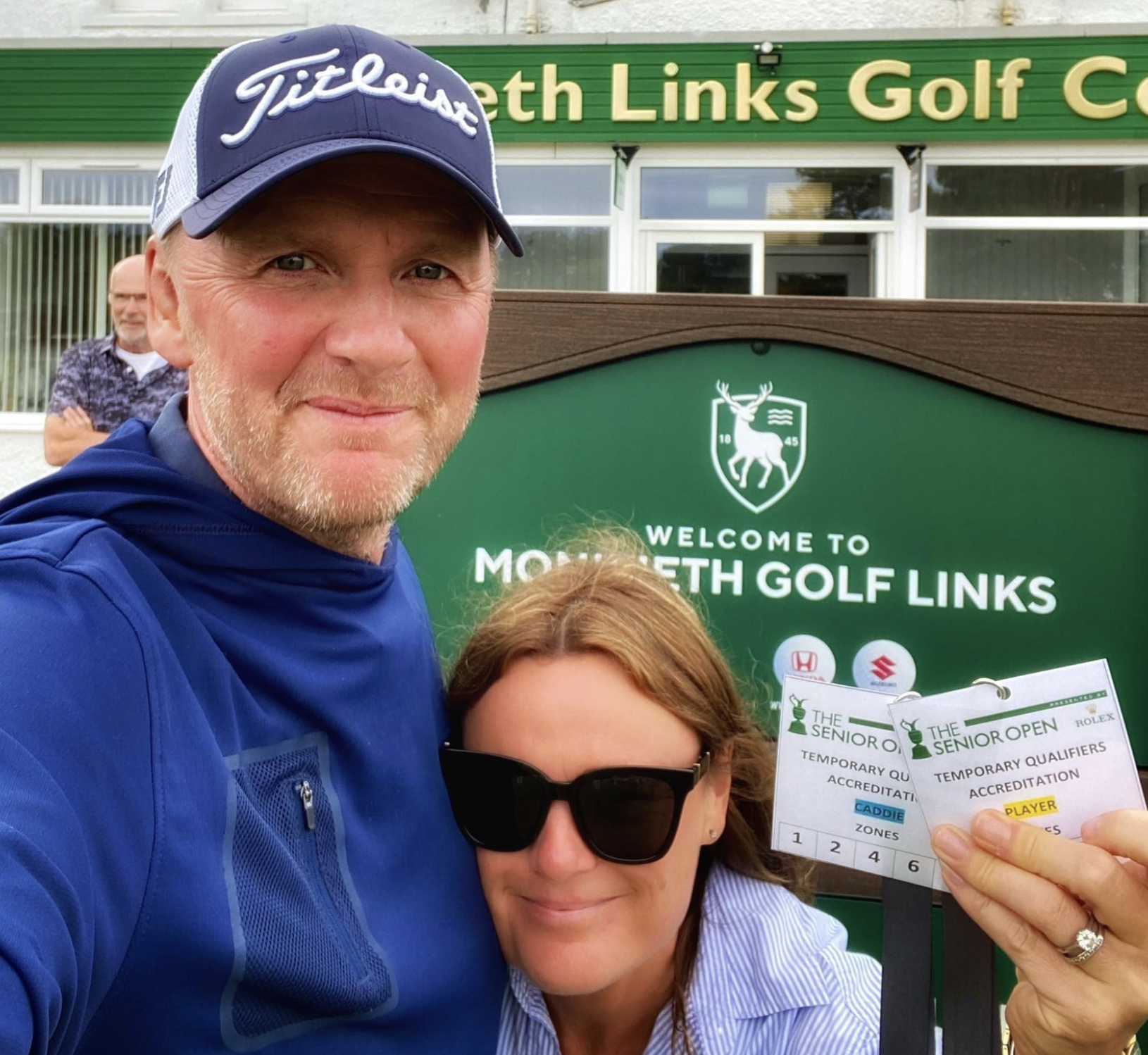 Two people taking a selfie outside of the North Links Golf Club. One person is a man wearing a blue Titleist cap and a blue jacket. The other is a woman with brown hair wearing black sunglasses, holding two golf tournament scorecards. In the backgrou