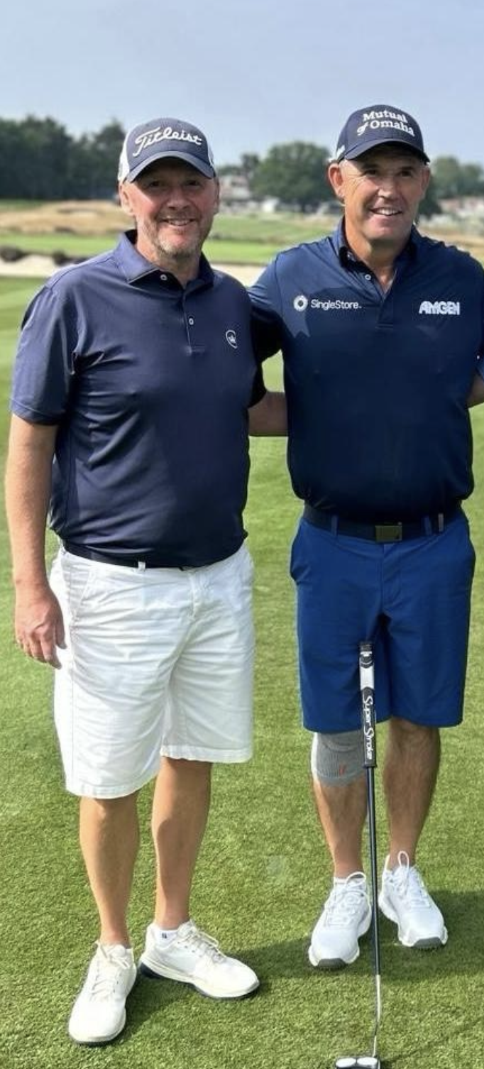Two men standing on a golf course, smiling. They are wearing golf attire and hats, with one holding a golf club.
