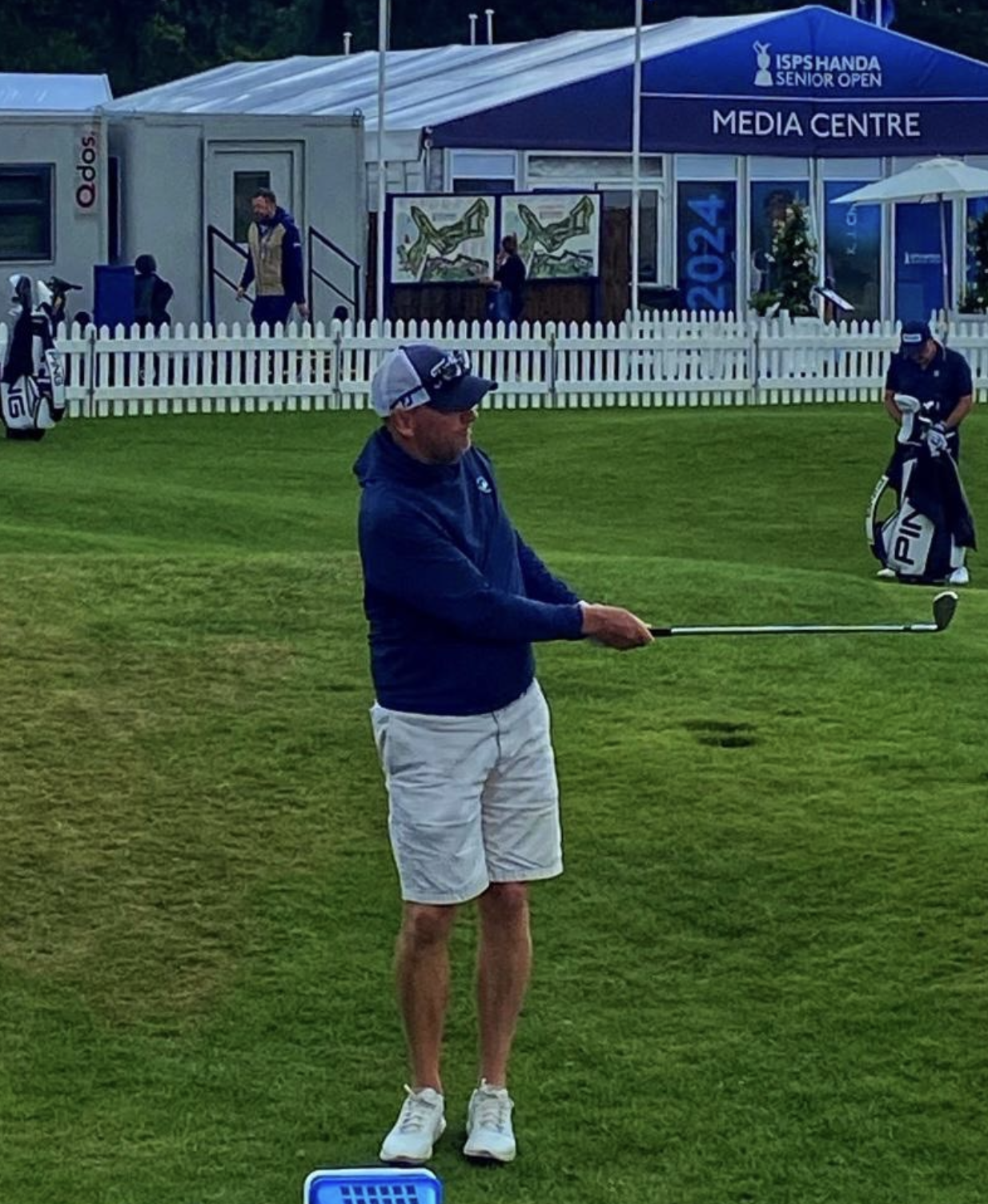 A man wearing a baseball cap, sunglasses, navy blue jacket, and beige shorts on a golf course preparing to hit a golf ball with a club.