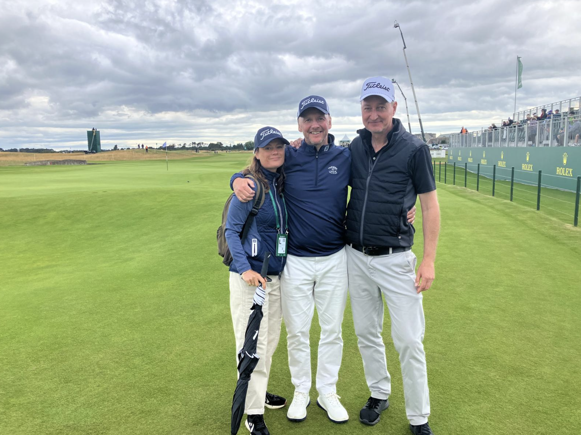 Three people standing on a golf course, smiling and posing for the photo, wearing golf caps and casual attire. The background features a flag on the green, spectators seated on the right side, and a cloudy sky.