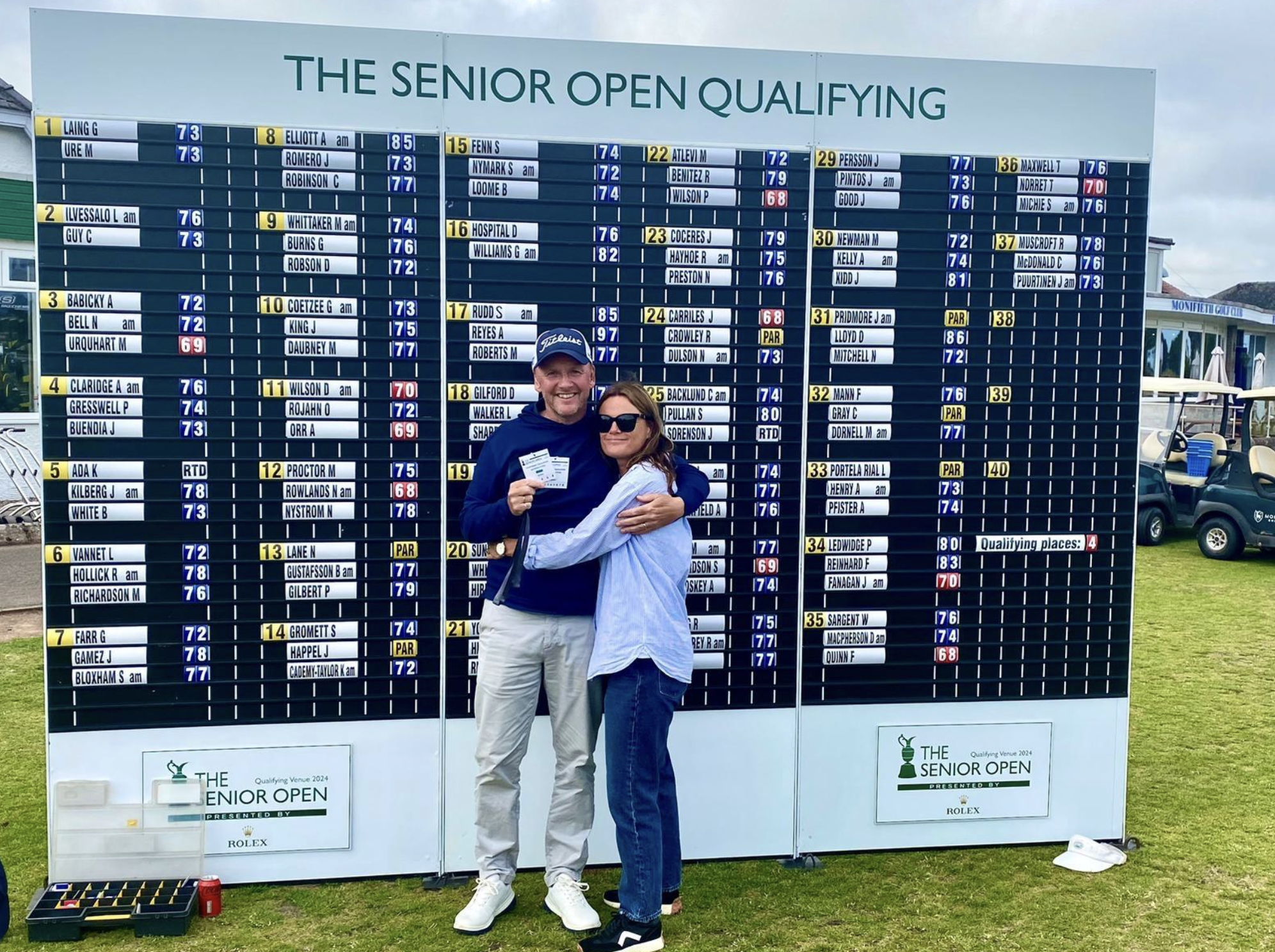 Two people smiling and hugging in front of a large board displaying the results of the Senior Open golf tournament.
