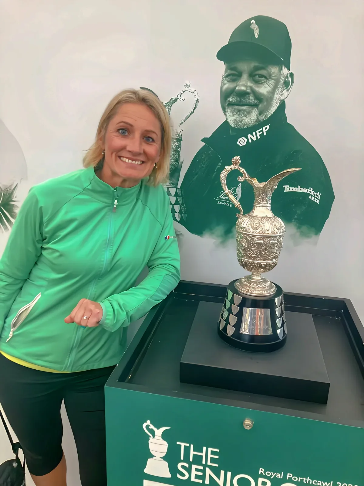A smiling woman in a bright green jacket stands next to a large, ornate silver trophy on a black base, which is placed on a green table with the logo and text 'The Seniors Royal Porthcawl 2023.' Behind her is a partially shading splashed background with a large black-and-white illustration of a man in a cap and sportswear, smiling, with a smaller trophy illustration behind him.