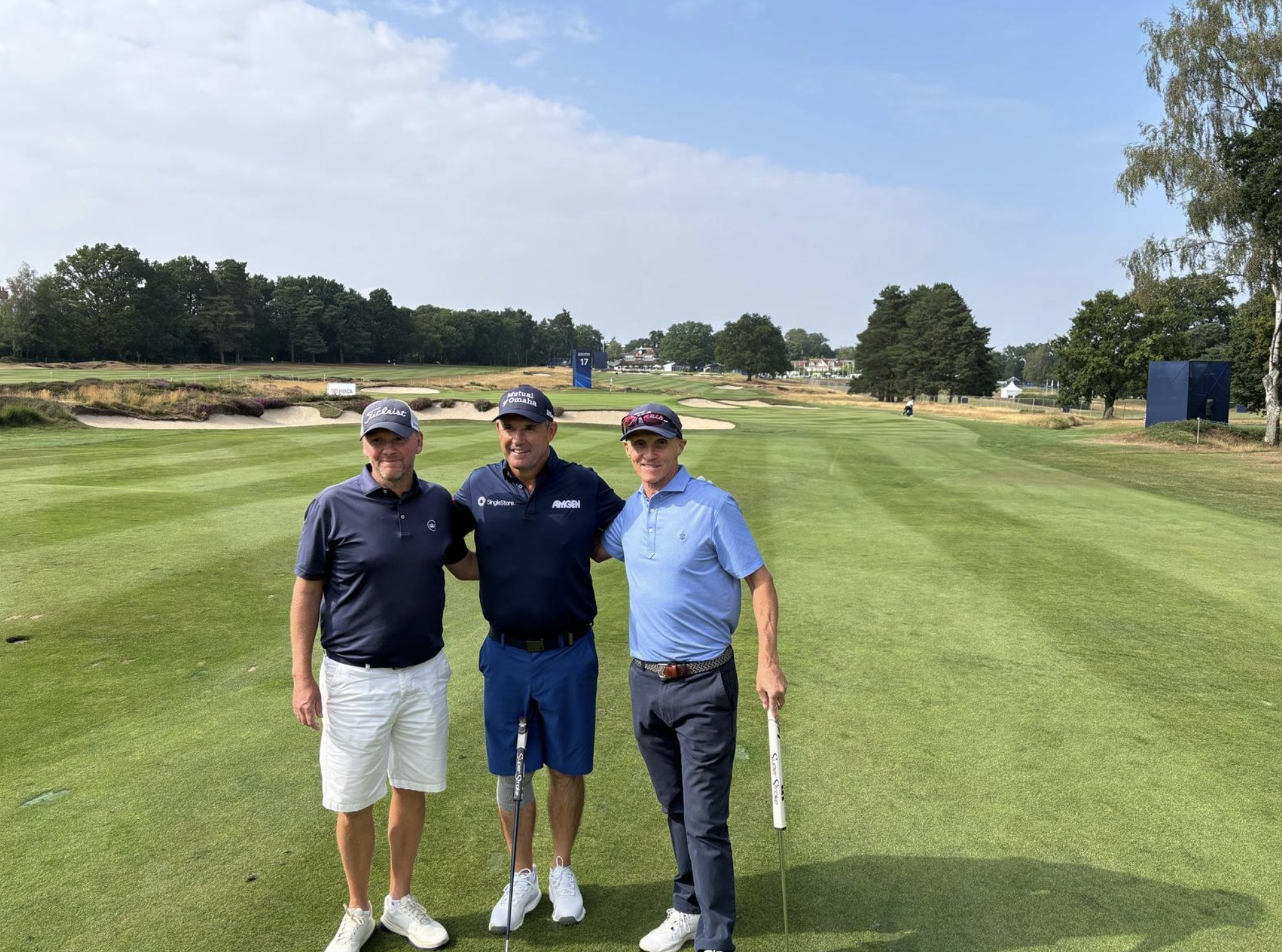 Three men standing on a golf course, smiling for the photo. They are dressed in golf attire and holding golf clubs, with trees, sand traps, and a scoreboard in the background under a partly cloudy sky.