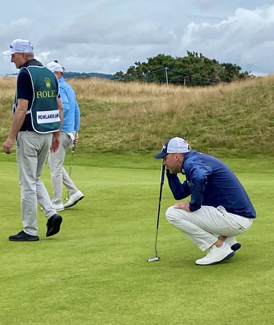 Man crouching on the golf green, holding a golf club to his forehead, with other men walking nearby on the golf course.