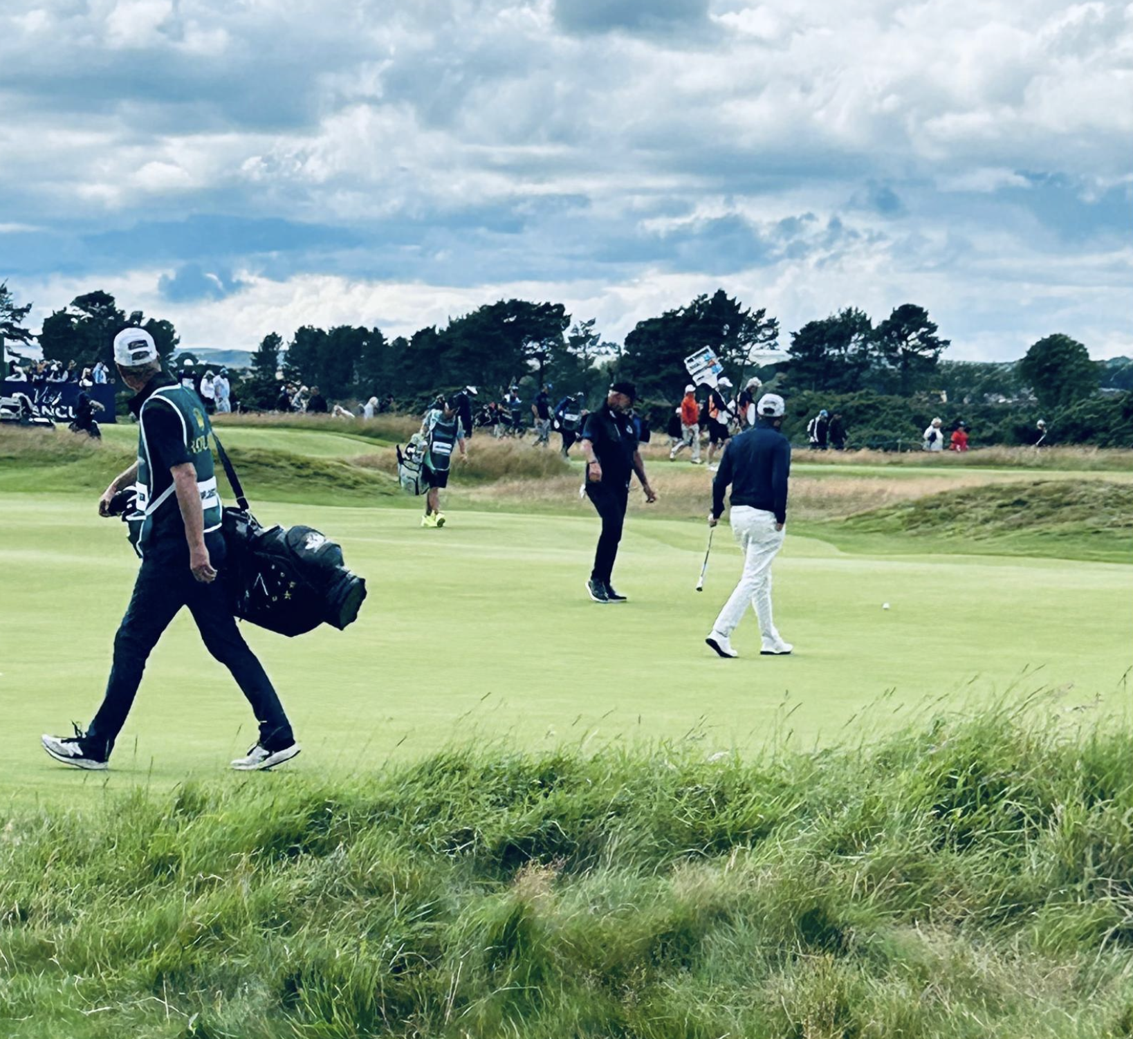 A group of people playing golf on a lush green golf course under a partly cloudy sky. Some are walking while others are preparing for their shot, with golf clubs and equipment visible.