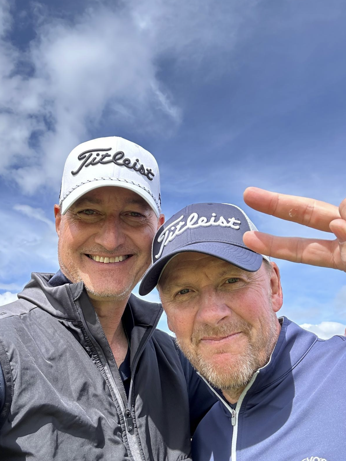 Two men taking a selfie outdoors, wearing Titleist caps and sports jackets, with a partly cloudy sky in the background. One man is smiling and making a peace sign.