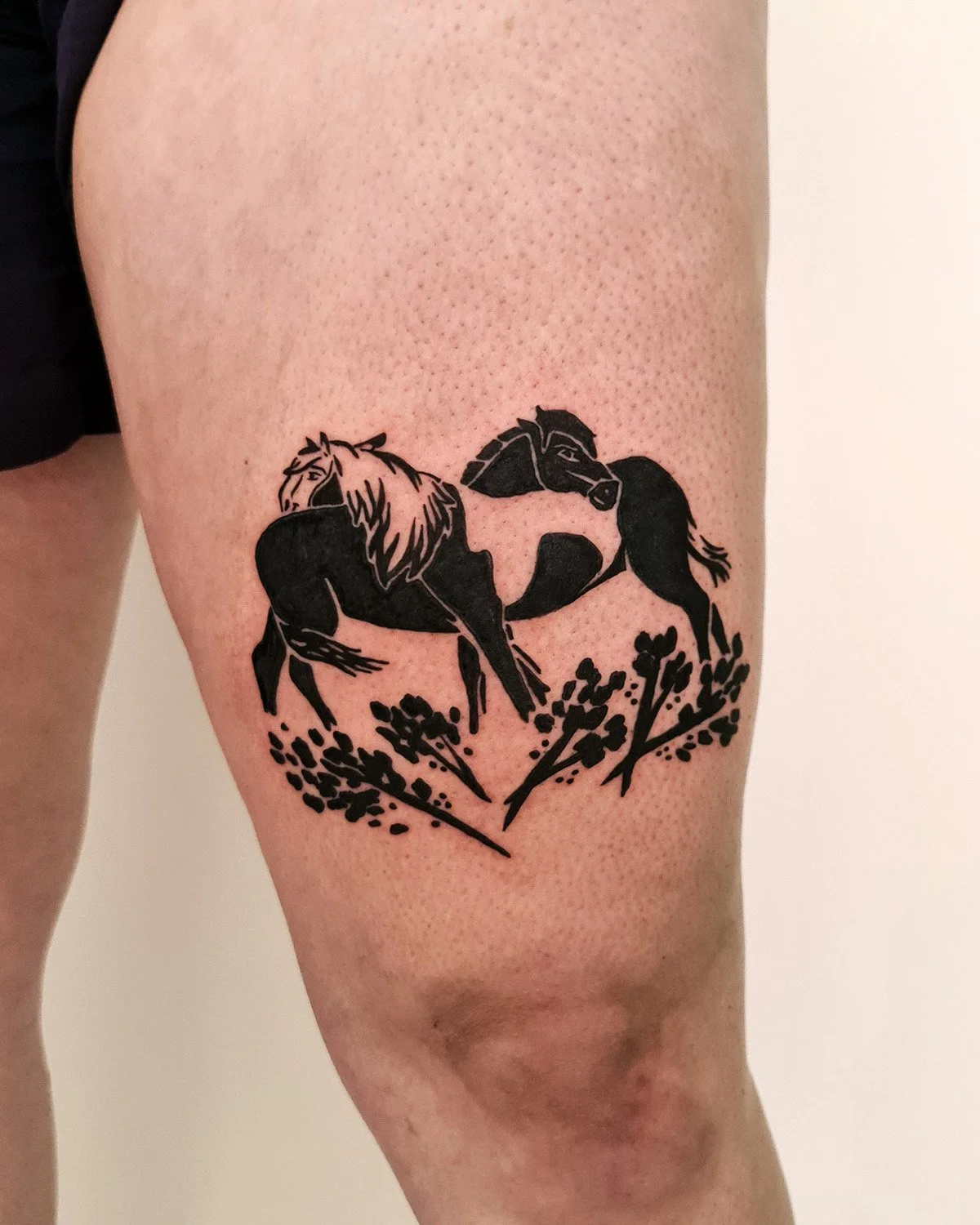 Tatouage de  Camille Tardif  de deux chevaux en silhouette noire, entourés de fleurs sur la cuisse.