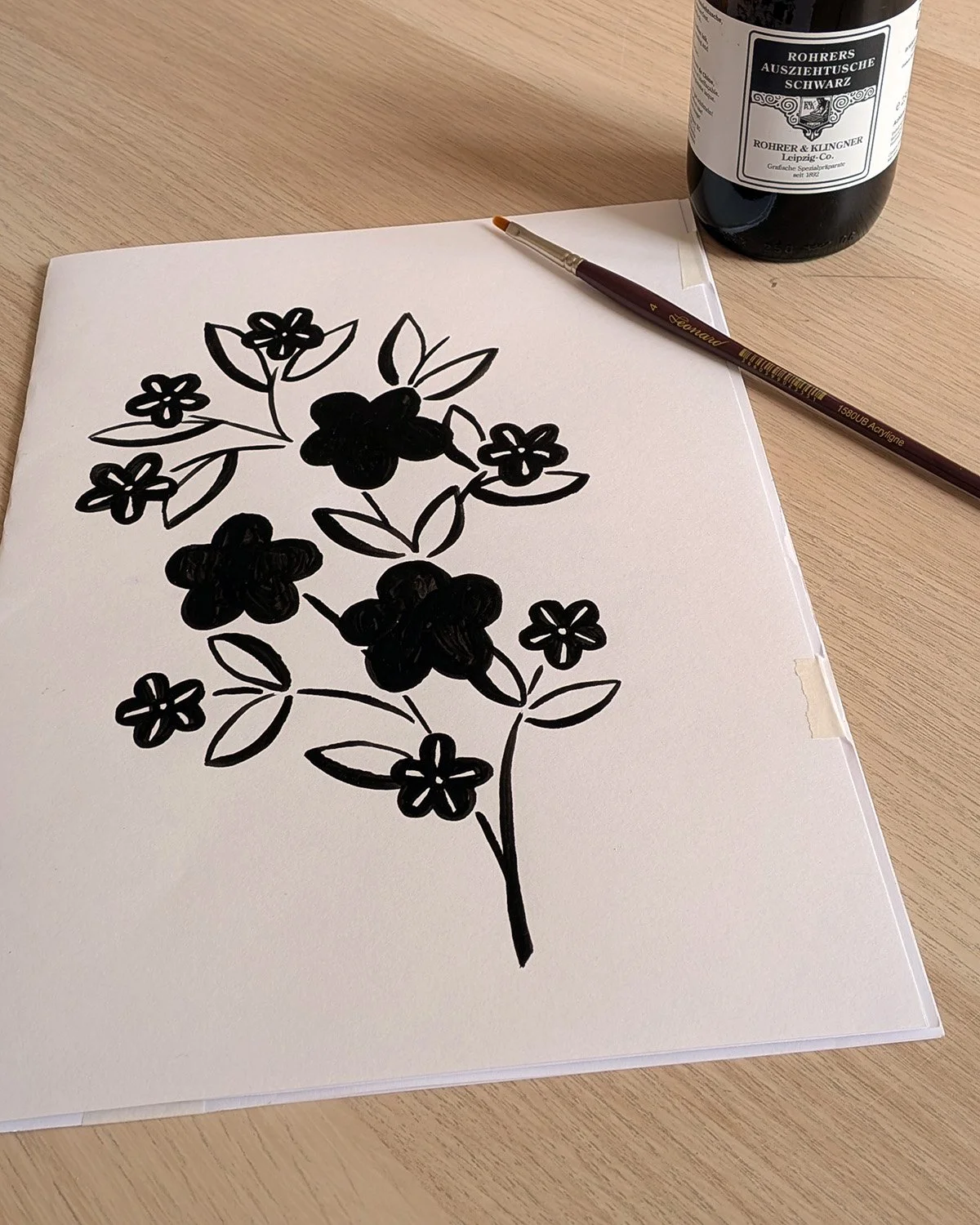 Dessins en noir de fleurs et de feuilles sur une feuille blanche, un pinceau et une bouteille de peinture à l'encre noire sur une surface en bois.