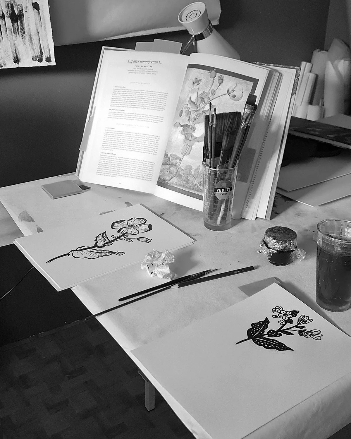 Une table d'art avec des illustrations de fleurs, des pinceaux, des peintures, un livre ouvert avec des images de nature, et divers matériaux pour la peinture.