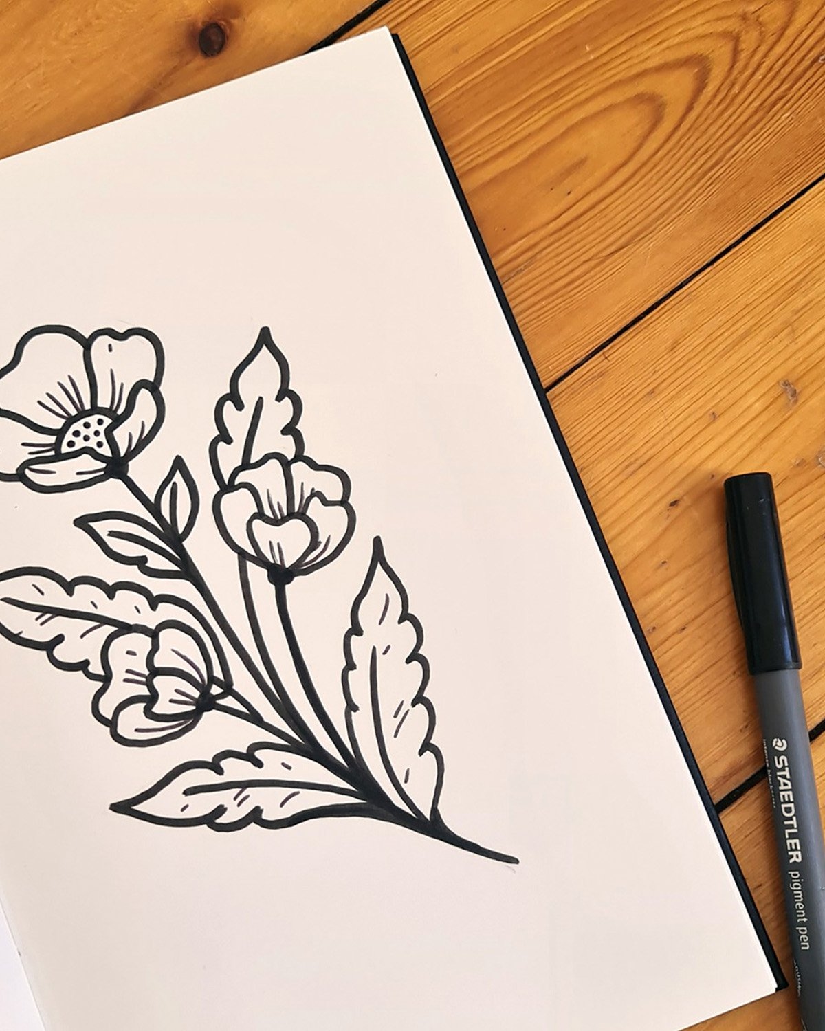 Dessin d'une branche de fleurs sans couleurs sur une feuille blanche, posée sur une surface en bois avec un marqueur à côté.