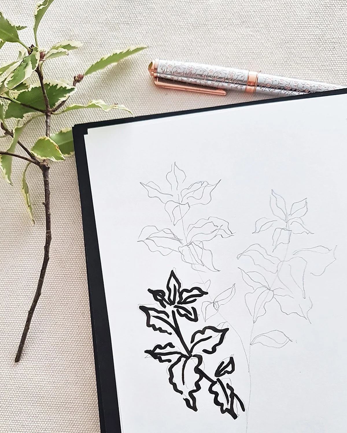 Croquis de branches de houx avec feuilles, un stylo à motif floral, un calepin sur une surface claire.