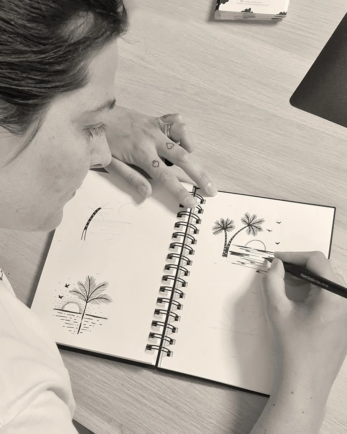 Une personne dessine une scène de plage avec des palmiers, un soleil couchant et des oiseaux dans un carnet.