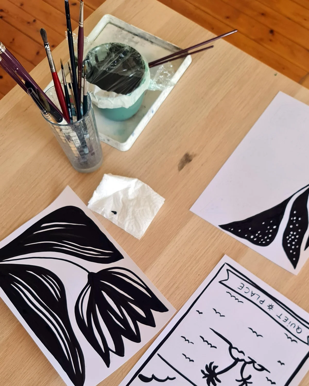 Table avec des œuvres d'art en noir et blanc, pinceaux, un gobelet contenant des pinceaux, un bloc de papier humide, une serviette en papier, et un dessin de paysage avec un palmier et des oiseaux.
