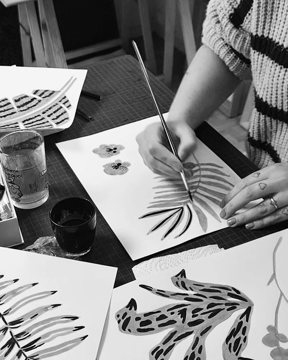 Une personne en train de peindre des motifs floraux et de feuilles sur une feuille de papier avec un pinceau, entourée de divers autres dessins et fournitures d'art.