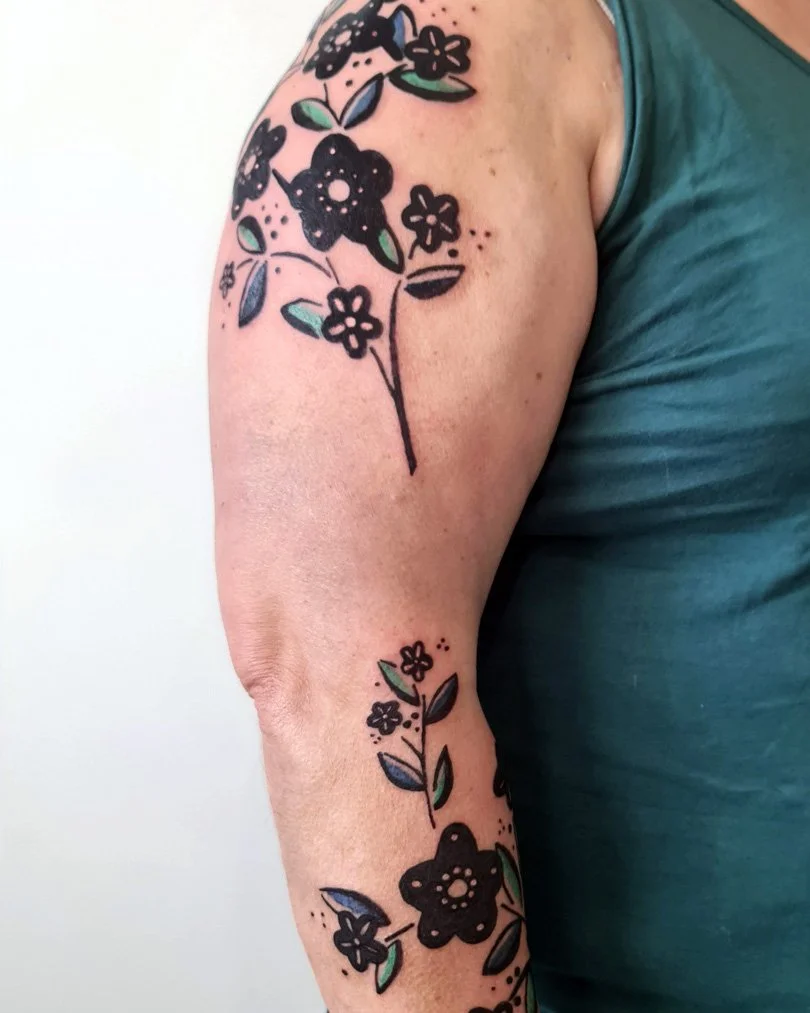 Tatouage de fleurs noires avec des touches de vert sur le bras d'une personne portant un t-shirt vert.