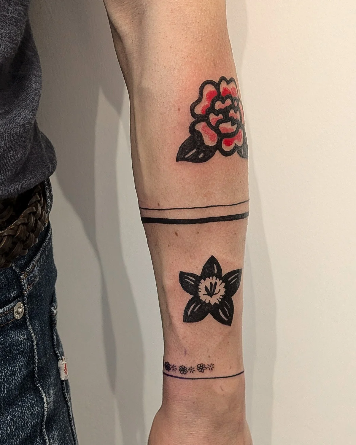 Tatouages sur le bras, incluant une rose rouge avec contours noirs, une fleur noire stylisée, des bandes noires et un motif floral discret.
