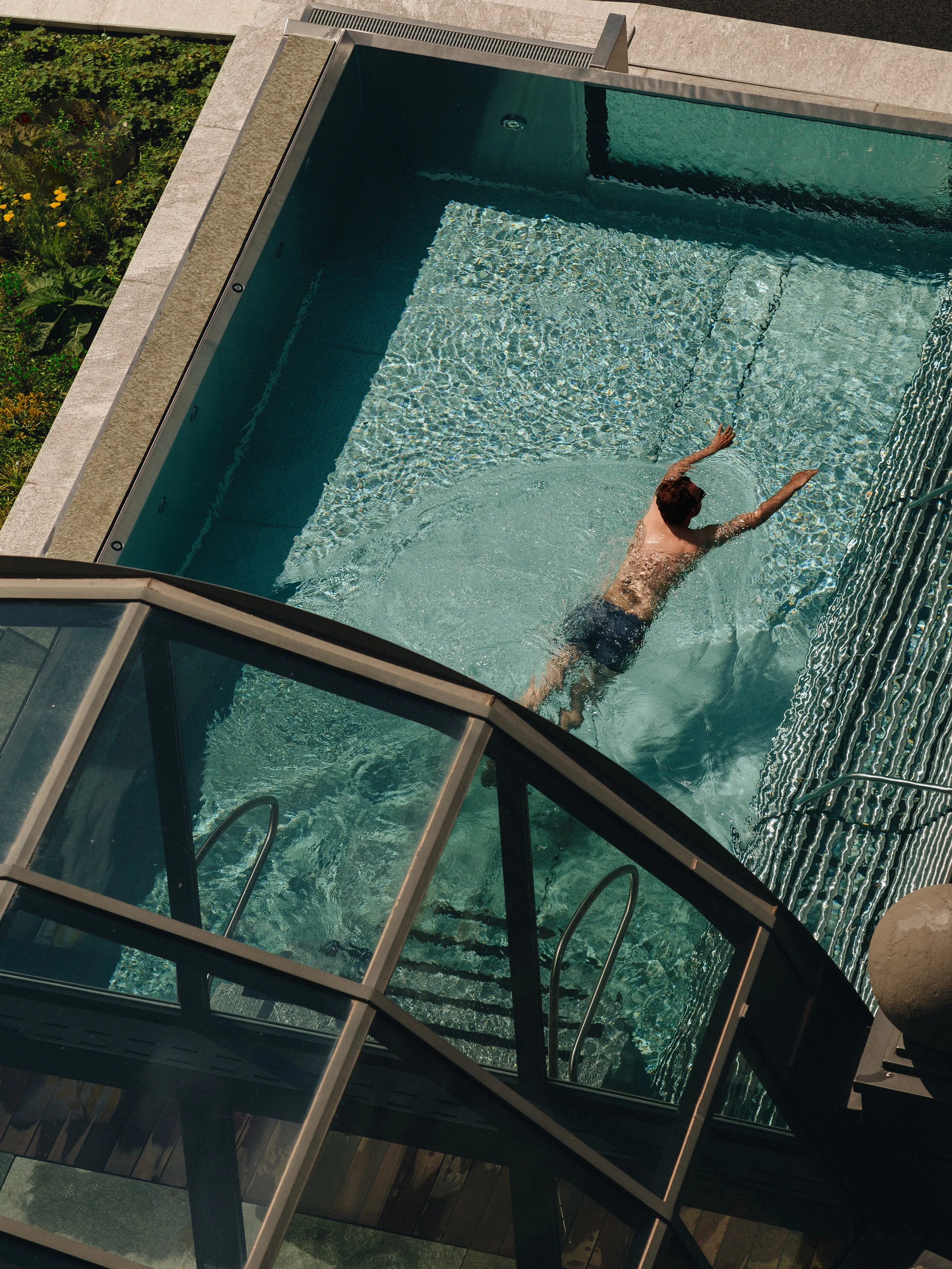 A person swimming in a backyard pool, viewed from above through a glass enclosure.