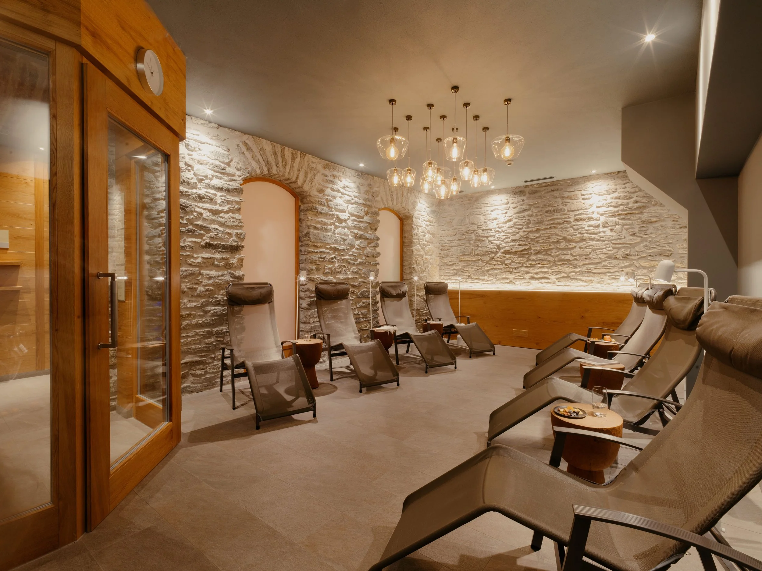 Spa or lounge area with beige reclining chairs, small side tables, a stone wall, hanging pendant lights, and a wood and stone interior design.