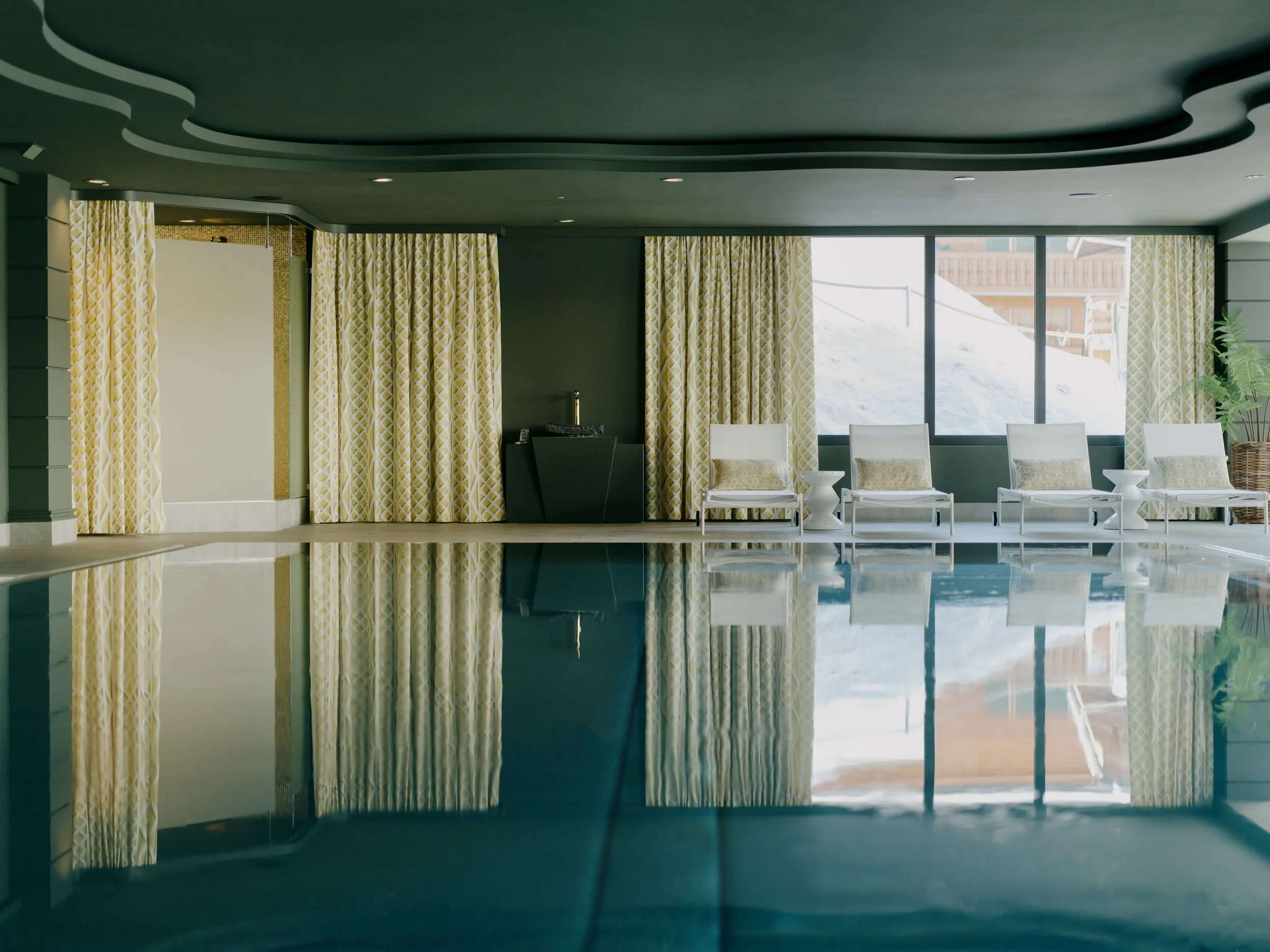 Interior of a room with yellow patterned curtains, white chairs, and large windows showing snow outside, with a swimming pool reflection in the foreground.
