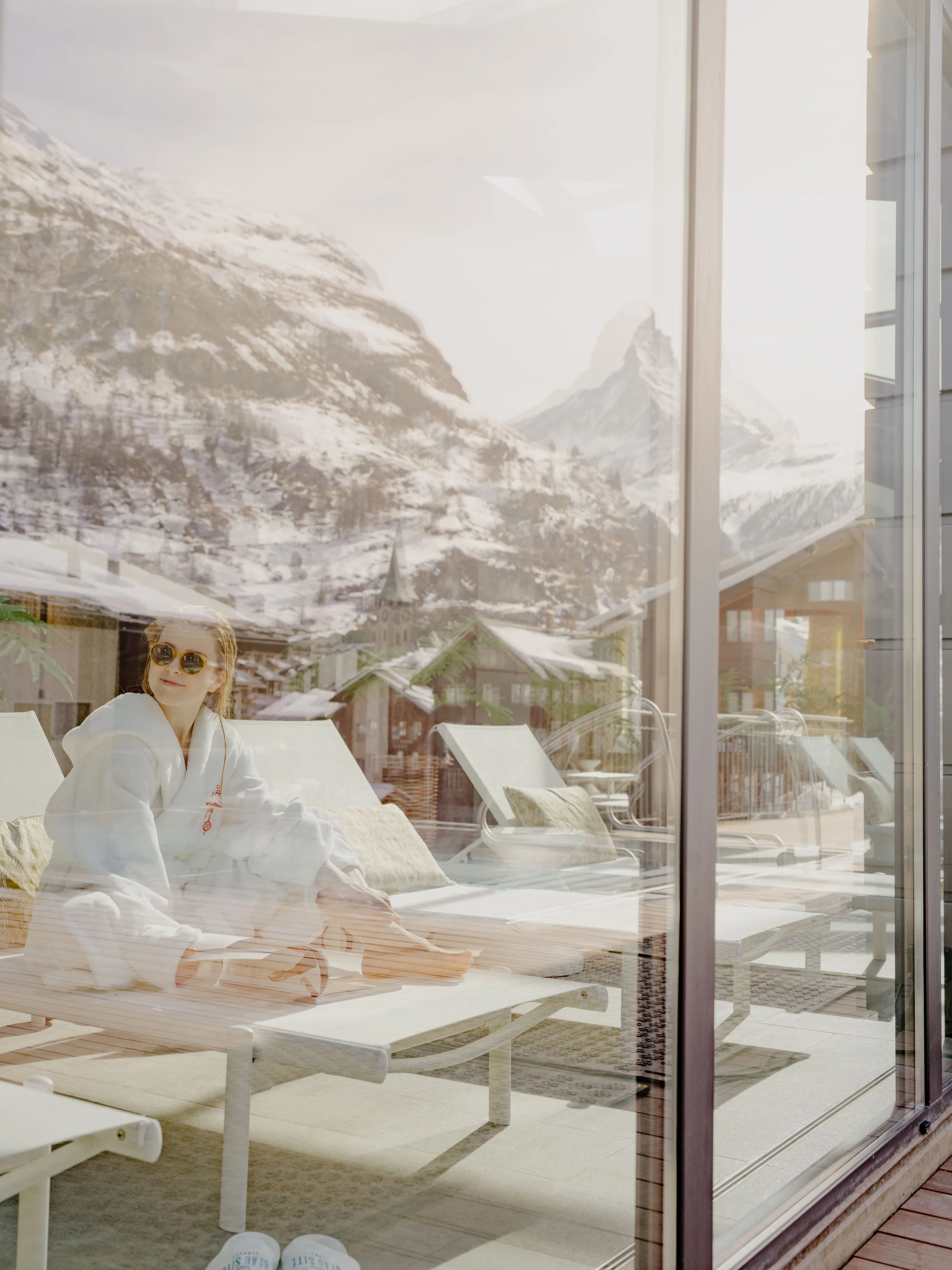 View through a glass window showing a woman relaxing outdoors on a lounge chair, wearing sunglasses and a white robe, with snow-capped mountains and a village in the background.