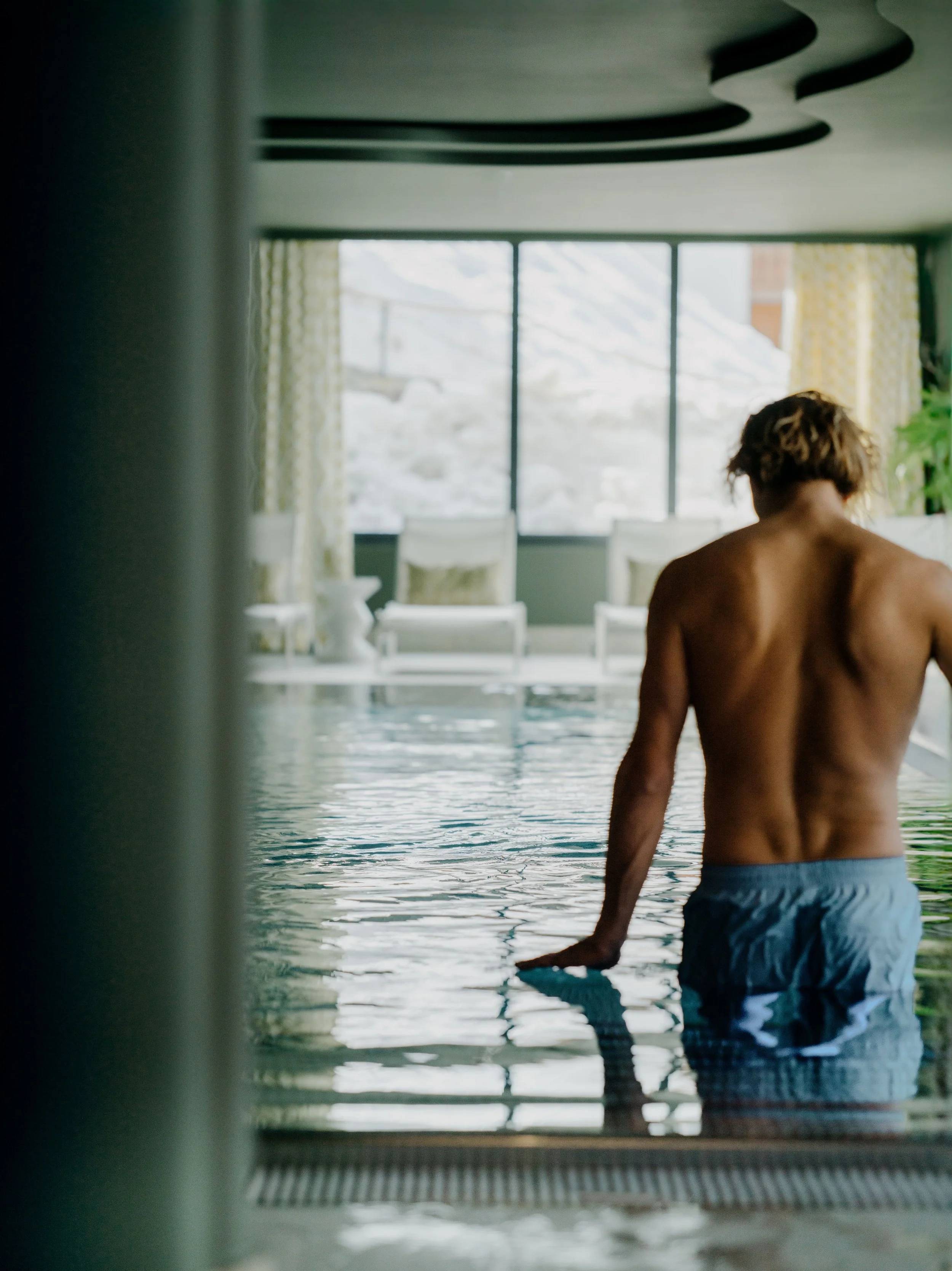 Person standing in a swimming pool, facing away, with large window behind showing outdoor scenery.