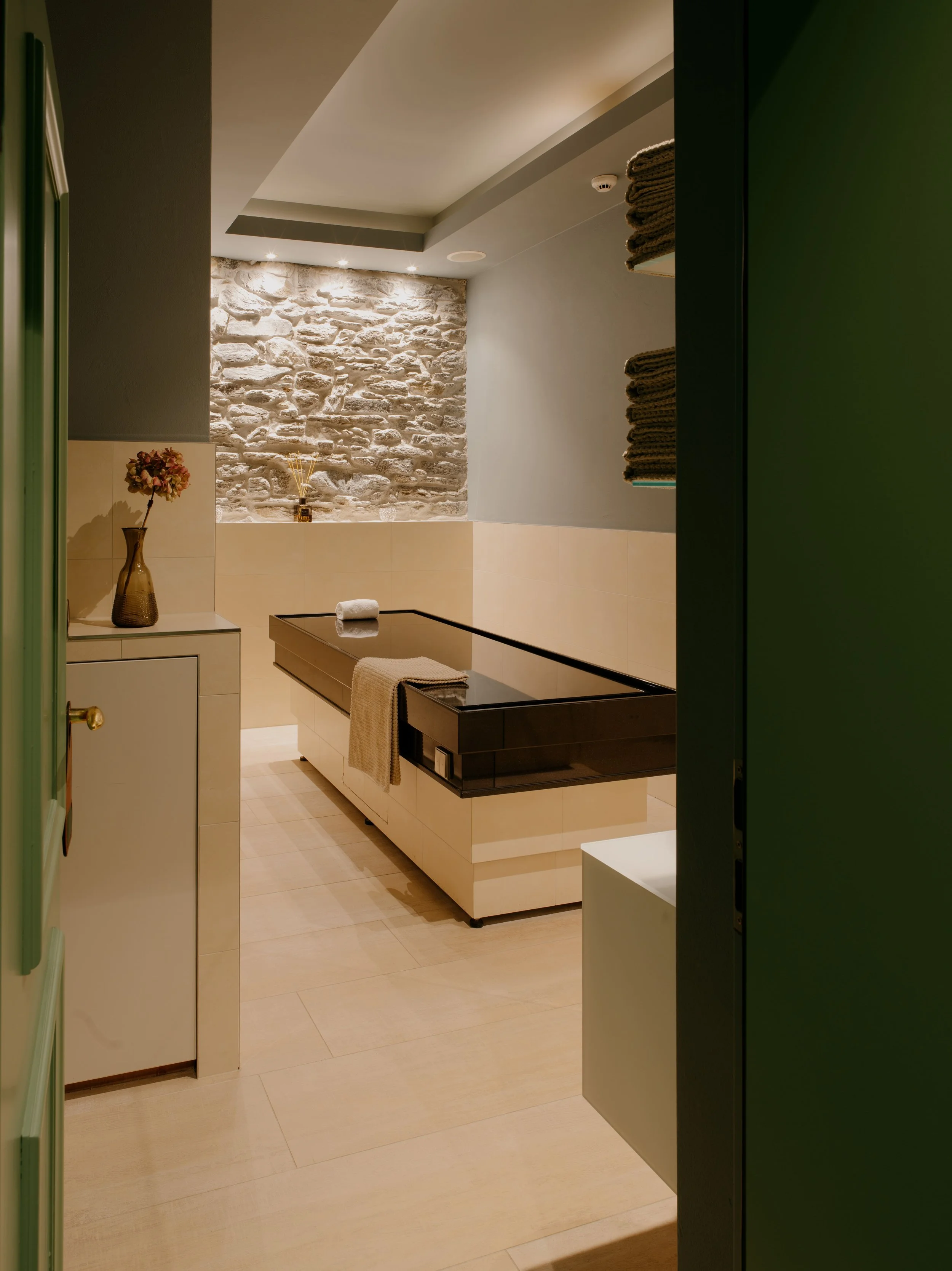 A spa or wellness room with a stone accent wall, beige tiled floor, dry towels stacked on shelves, a small white cabinet with a vase of flowers, a black bench with a towel, and soft lighting.