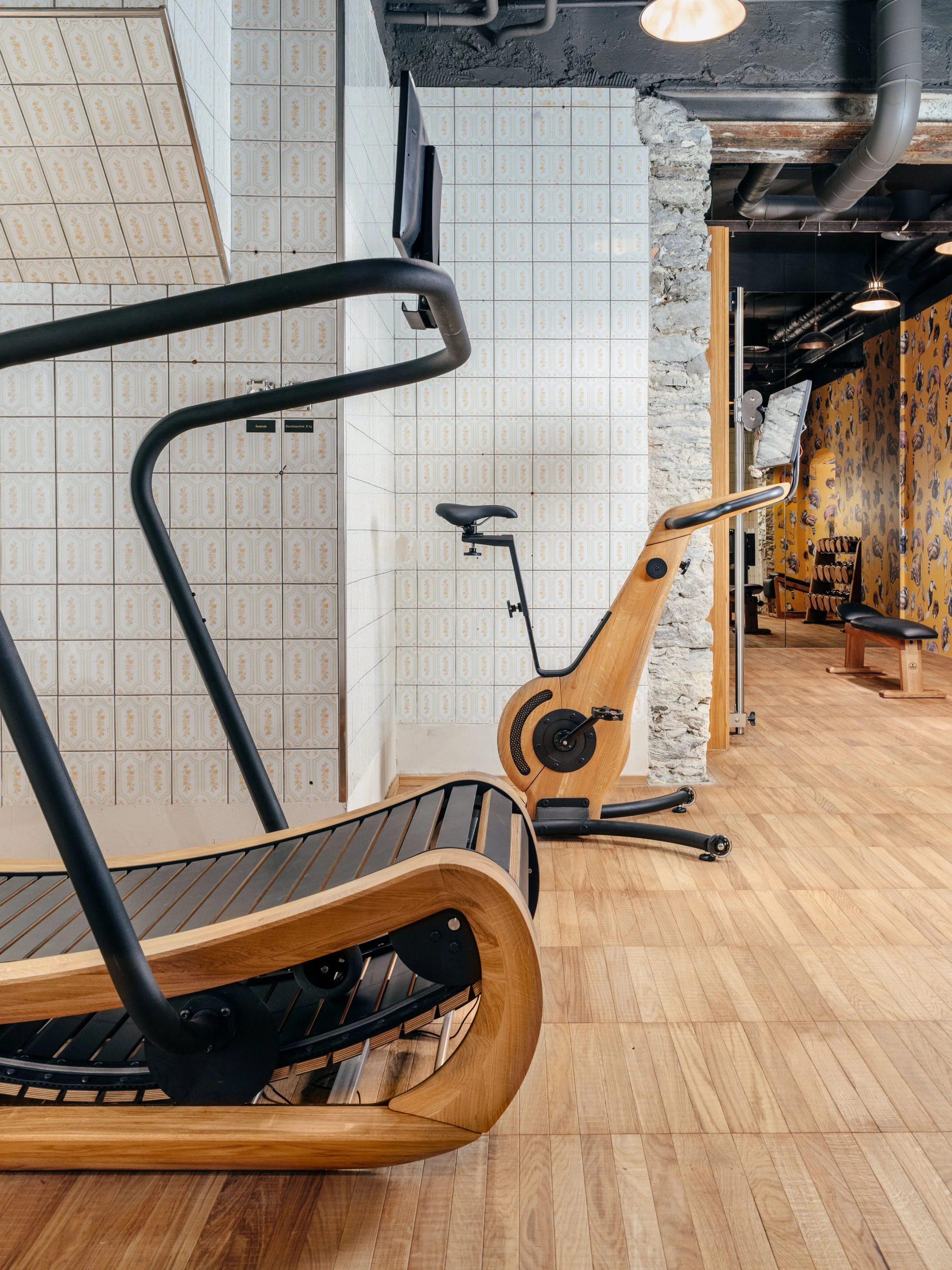 A modern workout space featuring a wooden elliptical trainer, a stationary bike, and part of a running treadmill. The room has wooden flooring, decorative tiled wall, and a combination of exposed brick and dark painted walls with industrial lighting.