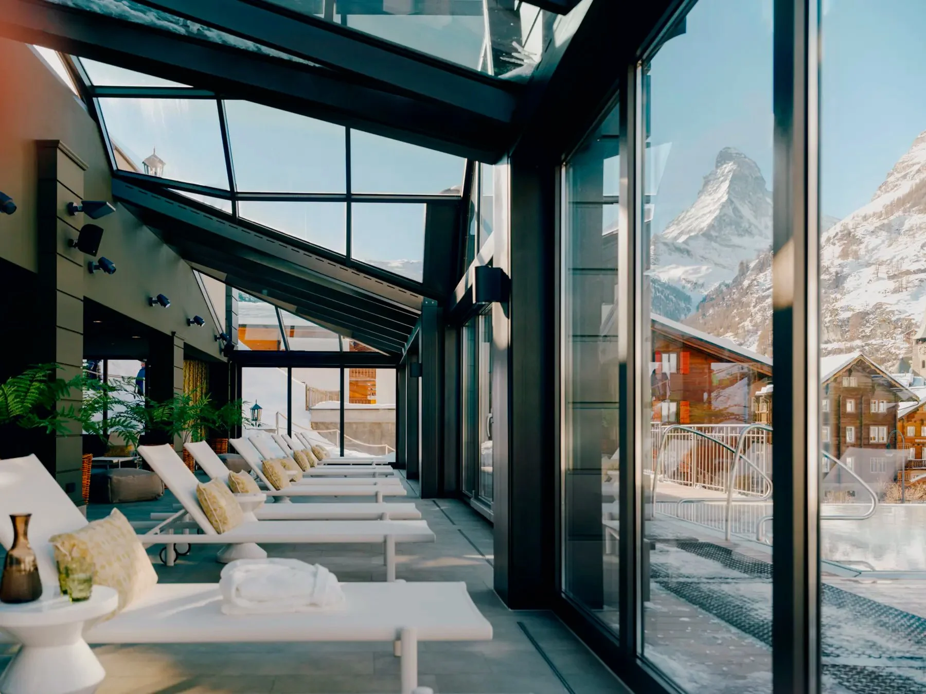 Indoor lounge area with white reclining chairs and beige pillows facing large glass windows showing snow-covered mountains in the background.