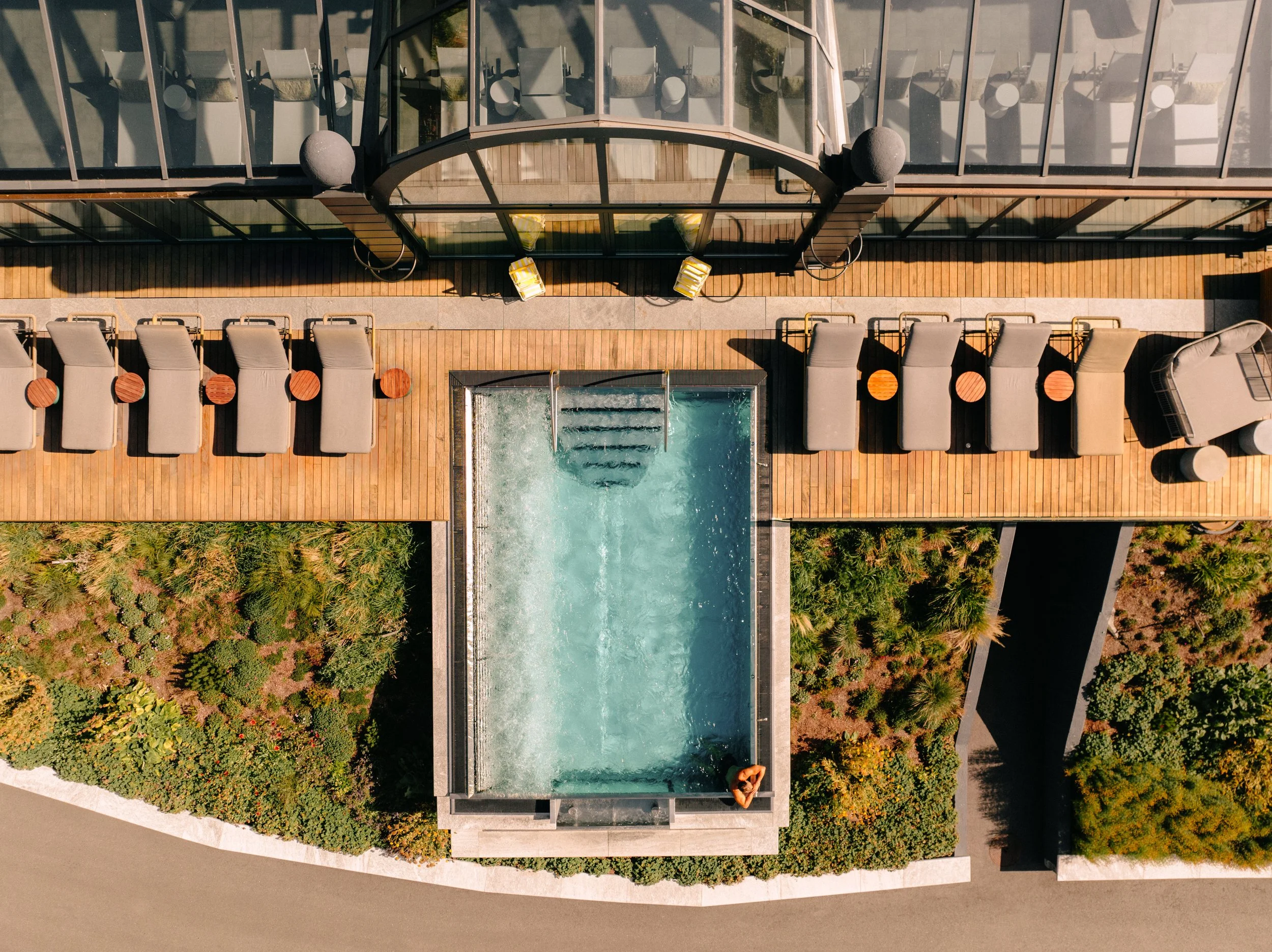 An aerial view of a modern building with a rooftop pool and deck area surrounded by lush greenery. There is one person lounging by the pool and several chairs and small tables on the deck.