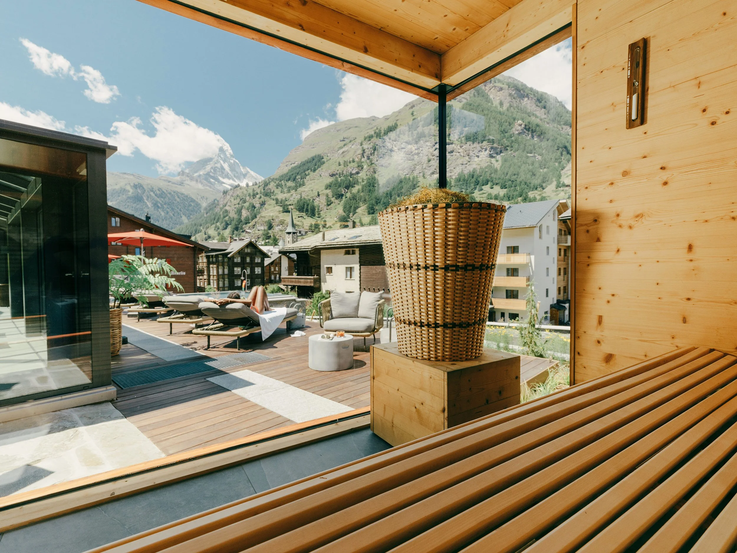 A balcony with wooden flooring and walls overlooking a scenic mountain village with a church steeple, surrounded by green hills and snow-capped mountains in the background.