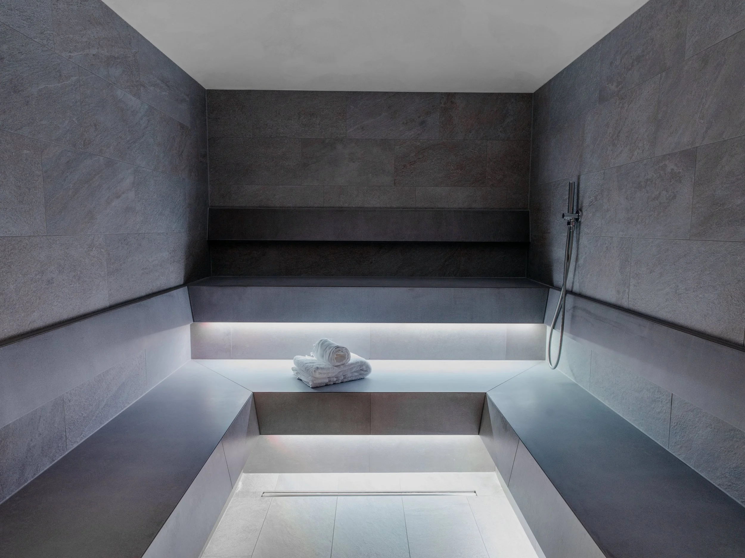 Modern shower area with grey stone tiles, a built-in bench, rolled white towels, and a handheld showerhead.