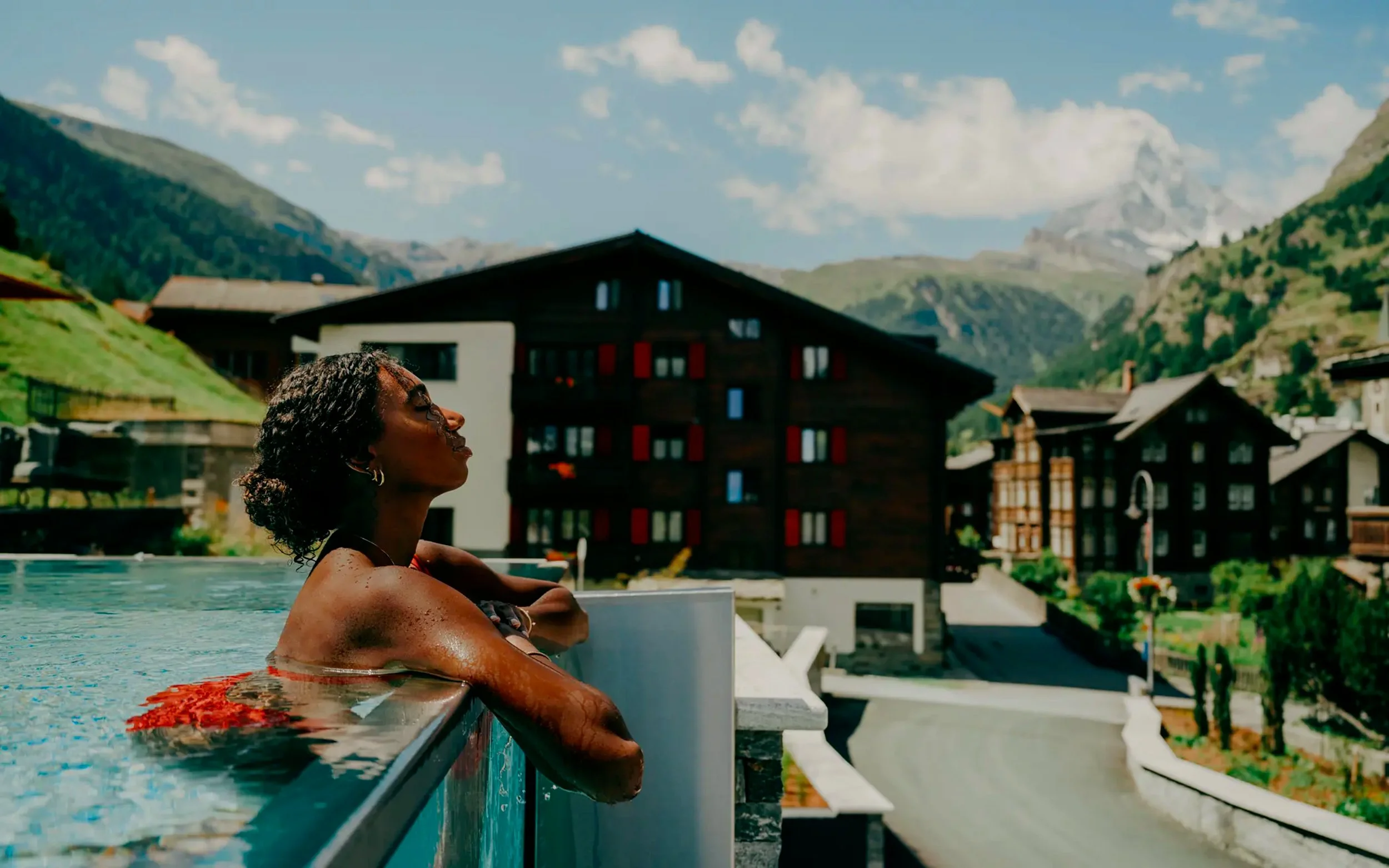 A woman relaxing in a swimming pool with a mountain village background on a sunny day.
