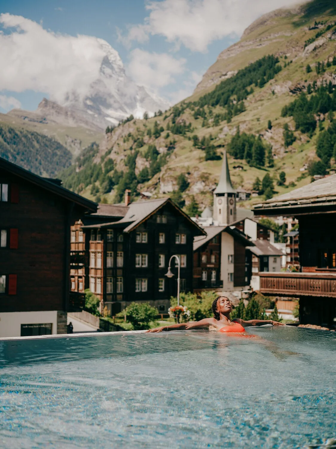A woman relaxing in an infinity pool with a scenic mountain village and snow-capped mountains in the background.