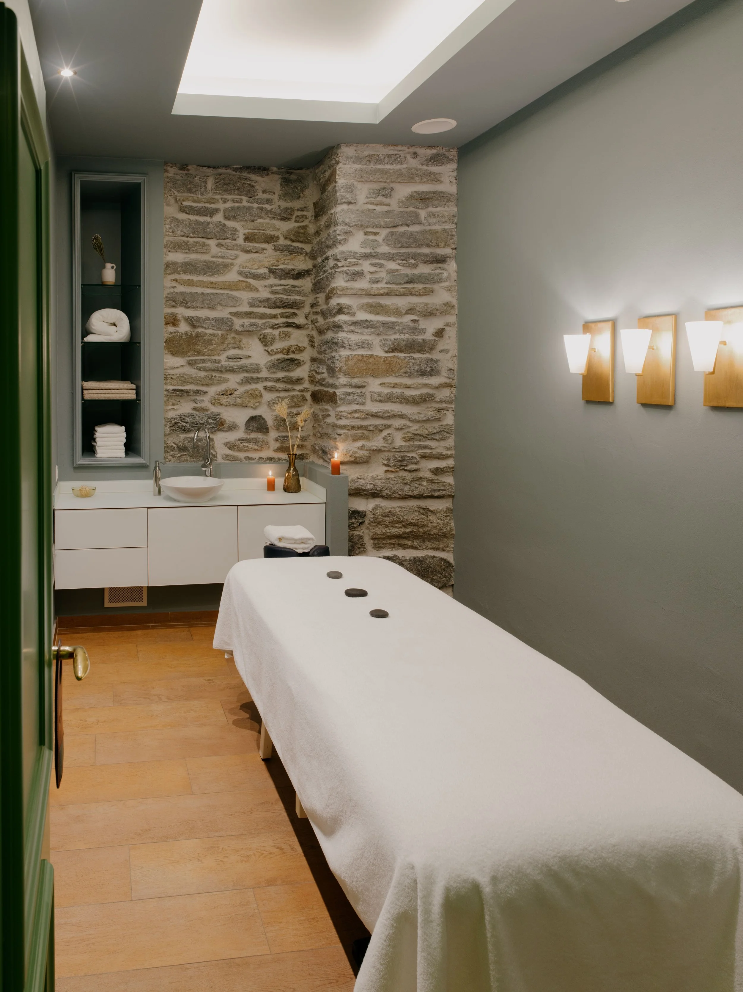 A massage table covered with a white sheet in a room with a stone wall. There is a small sink with a faucet and candles on the edge of the sink. The room has wooden flooring, a green wall, and a built-in shelf with rolled towels and decorative object