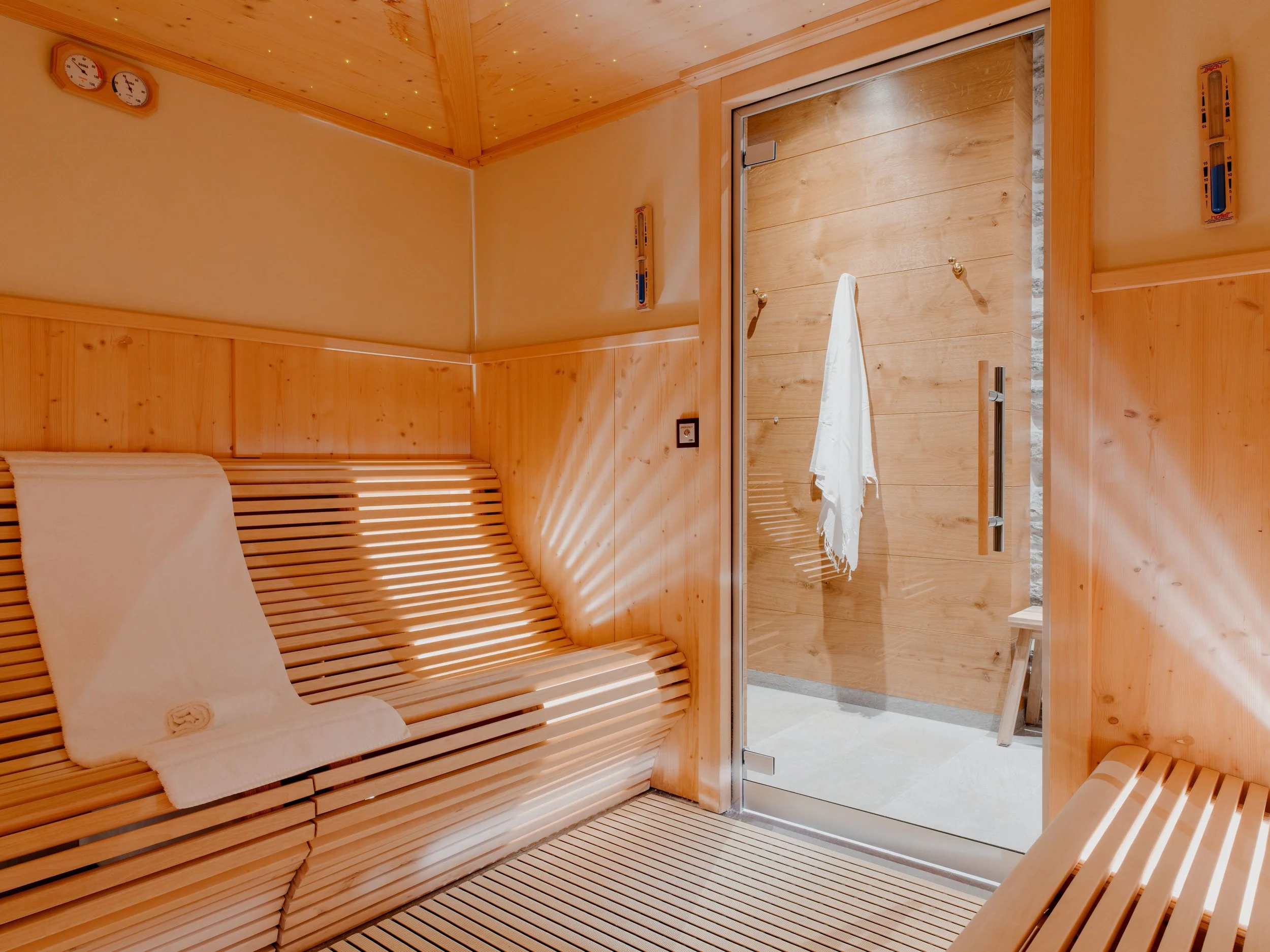 Interior of a wooden sauna with benches, thermometers, and a glass door leading to a shower area.