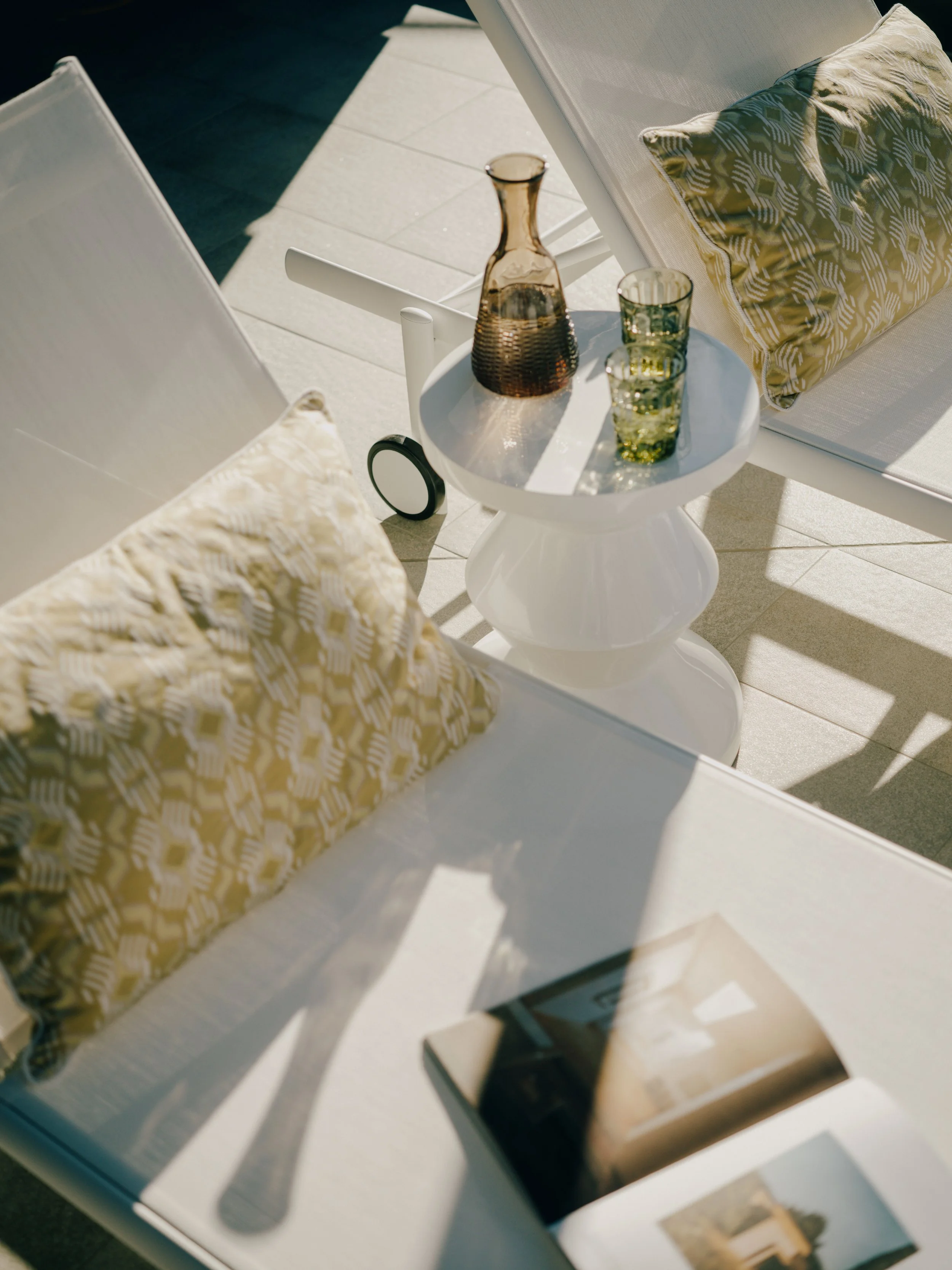 A white outdoor chair with a patterned cushion, a side table with a decorative vase, three glasses, and a watch, all illuminated by sunlight.