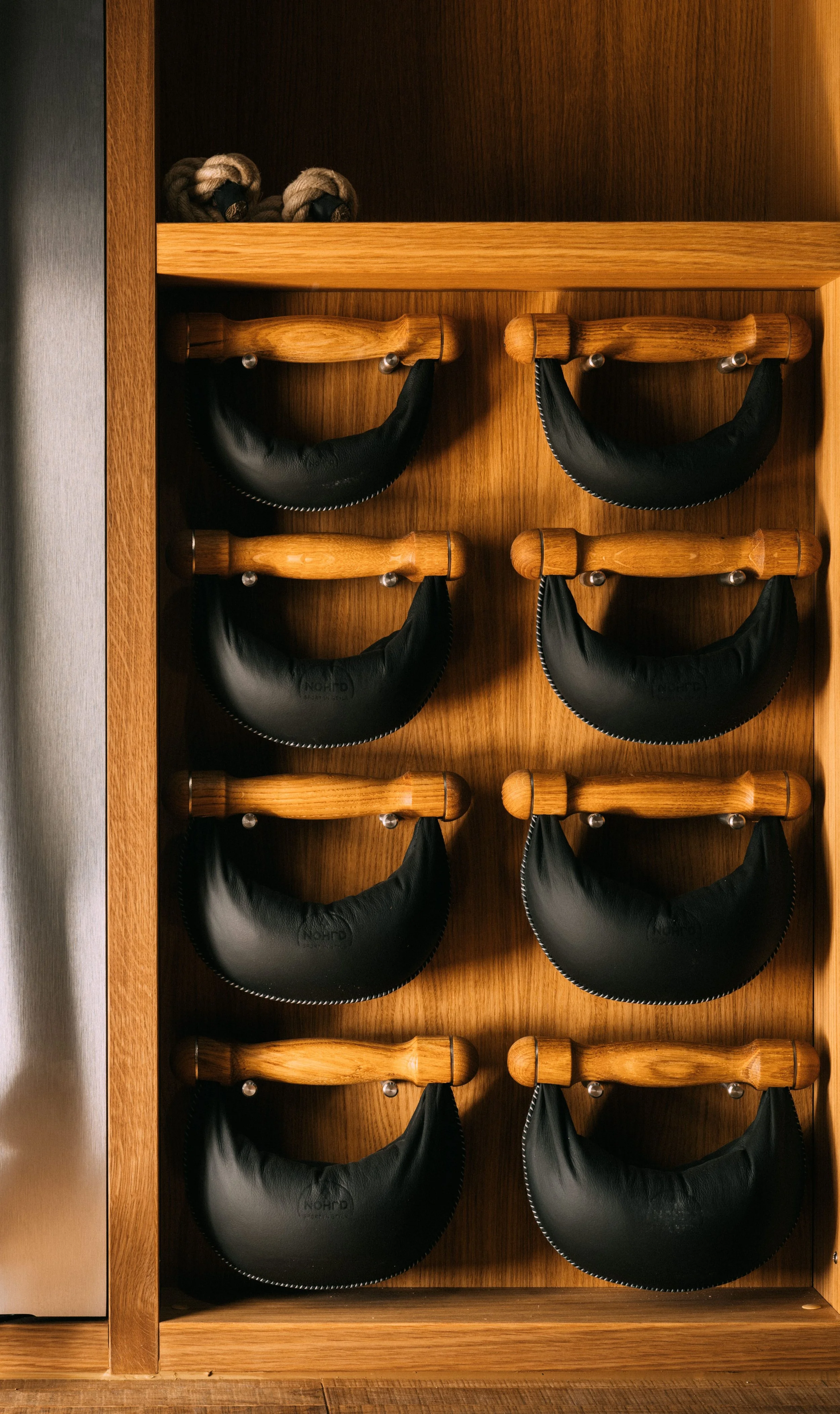 A wooden kitchen or storage cabinet with eight black leather potholders hanging on wooden hooks, arranged in two columns and four rows, with a small shelf at the top holding two pairs of braided oven mitts or gloves.