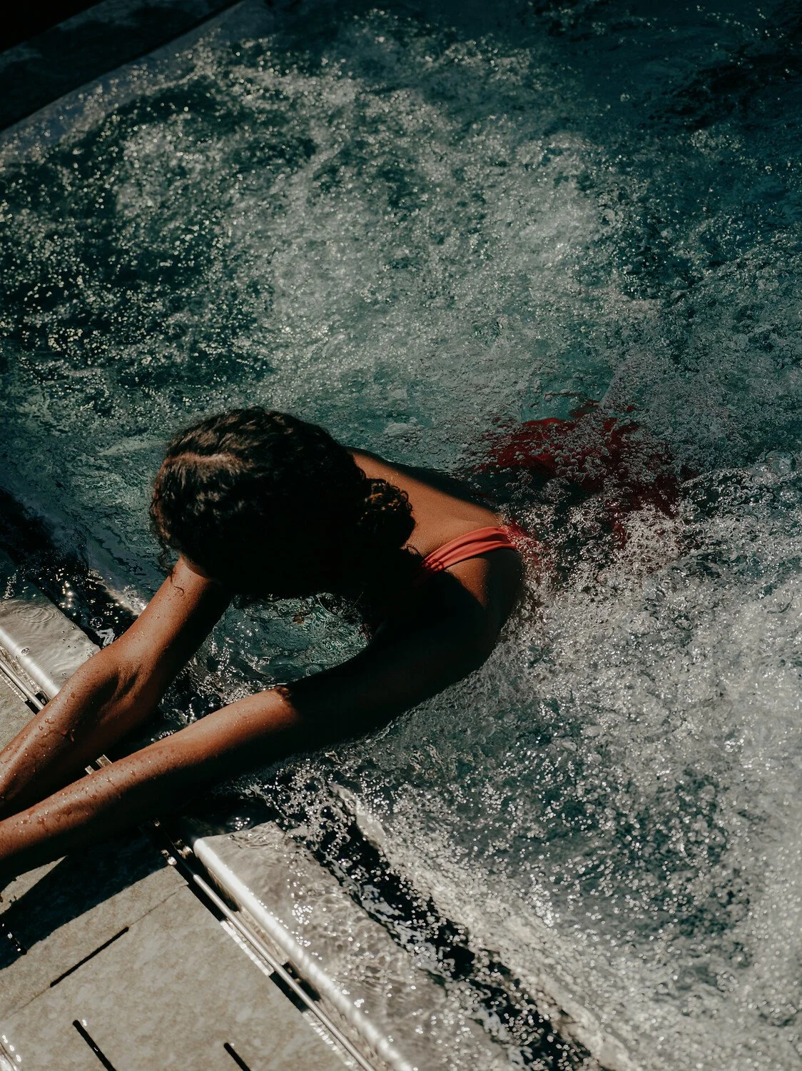 Person with curly hair wearing a red swimsuit leaning on the edge of a swimming pool with water splashing around.