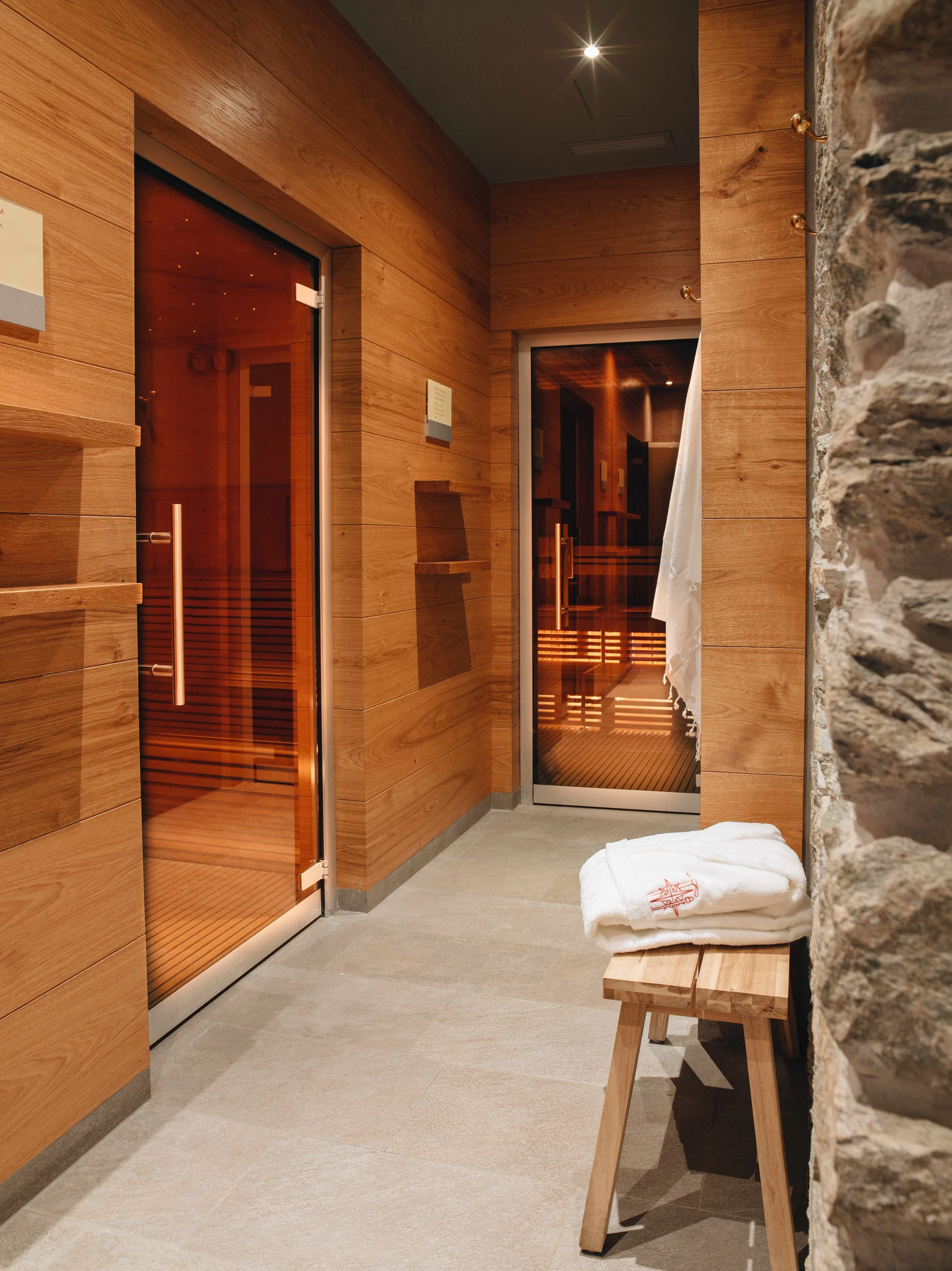 A cozy spa room with wooden walls and doors, stone wall on the right, beige tile floor, a white towel on a small wooden bench, and a window with a curtain.