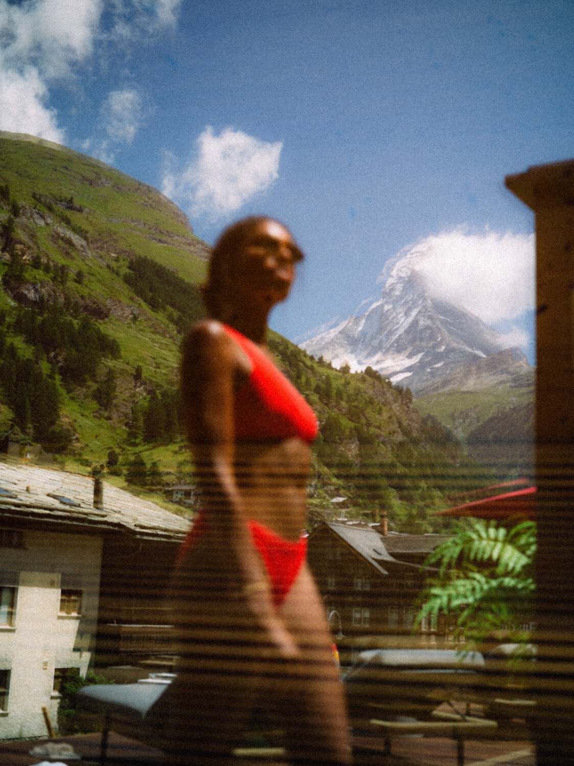 A woman in a red bikini standing outdoors with a mountain in the background, possibly in a rural area with houses and greenery.