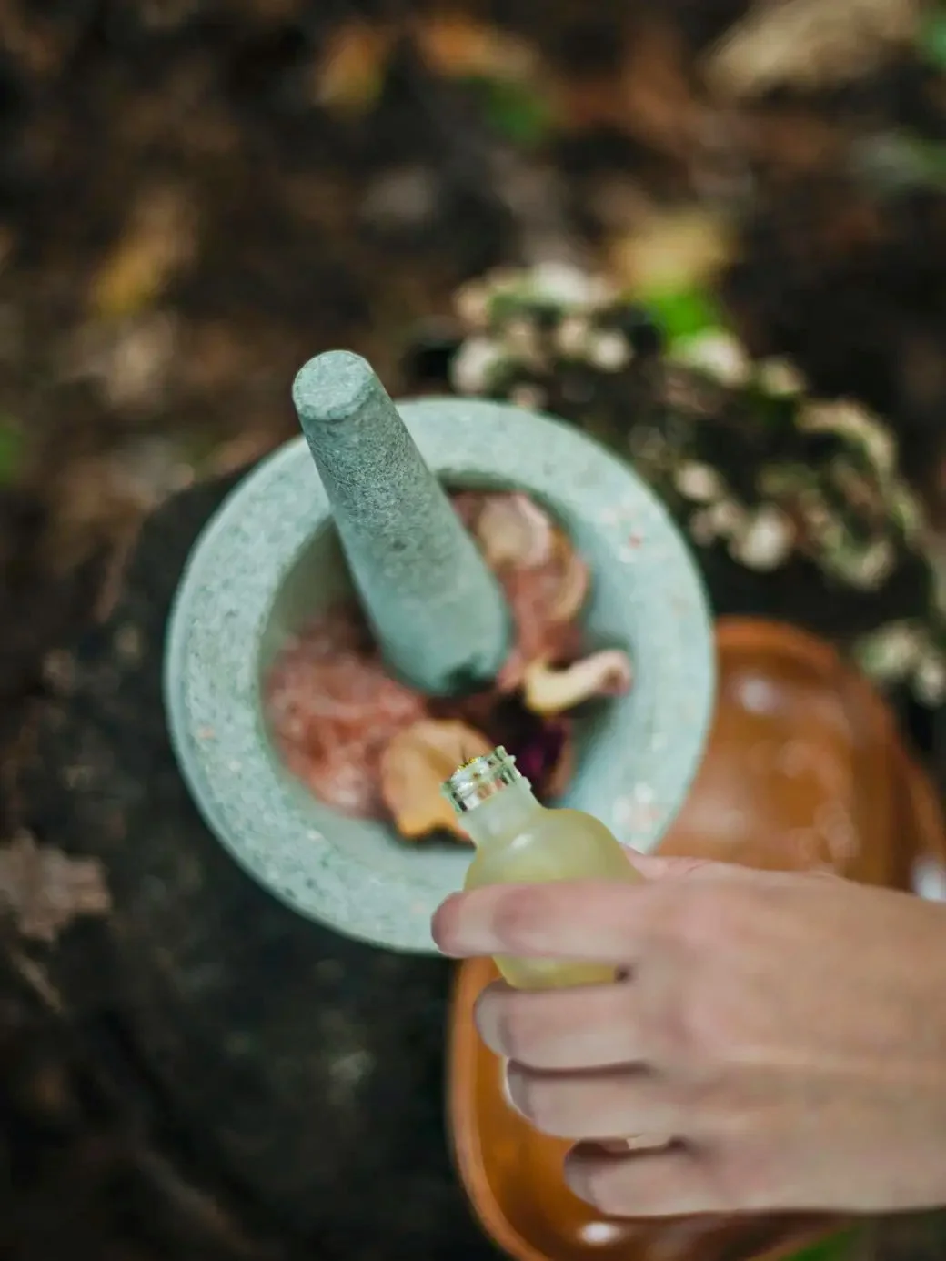 A person holding a small glass bottle near a mortar and pestle with herbs and dried flowers, likely preparing herbal medicine or tea.