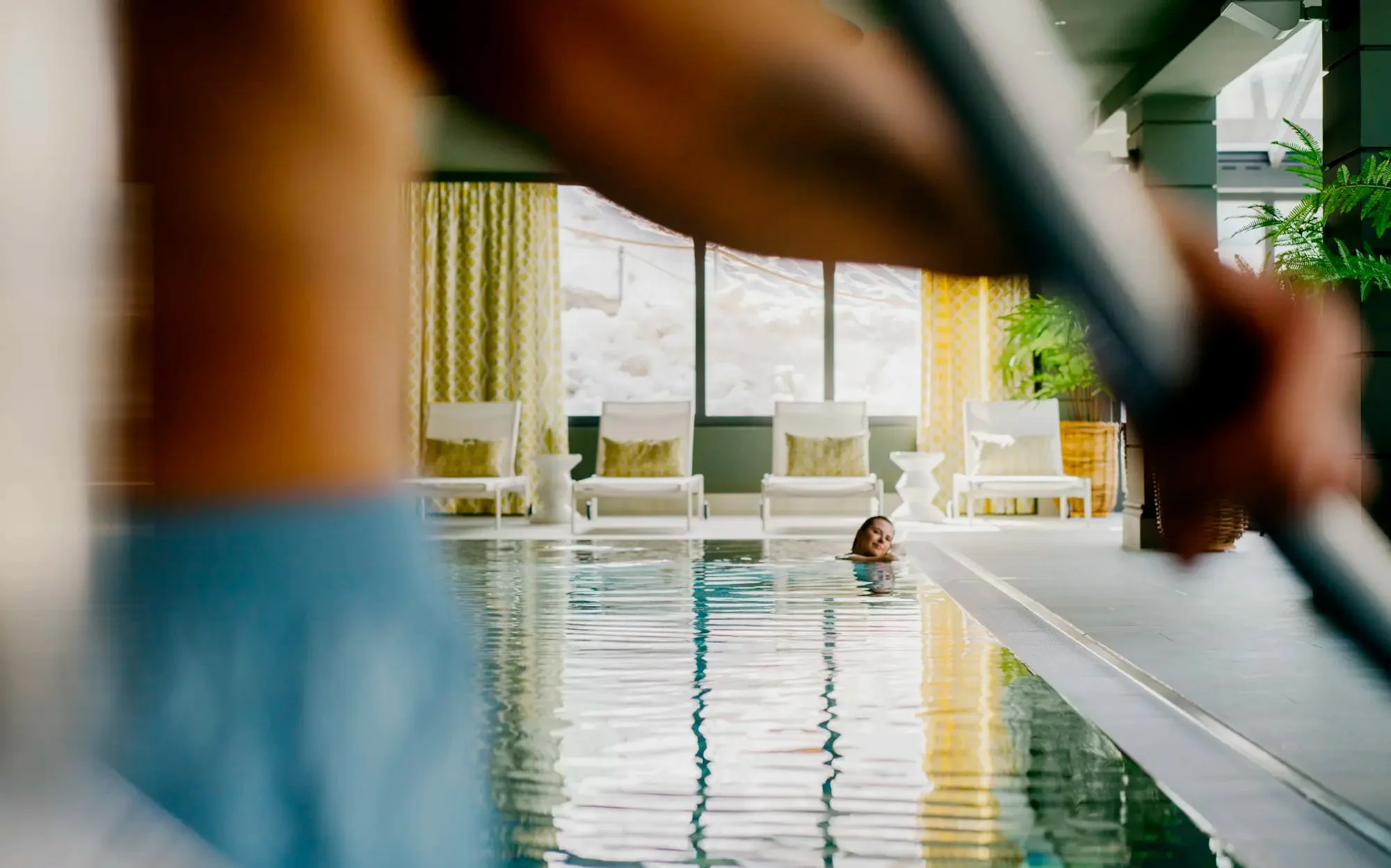 Person swimming in an indoor pool with chairs, plants, and large windows in the background.