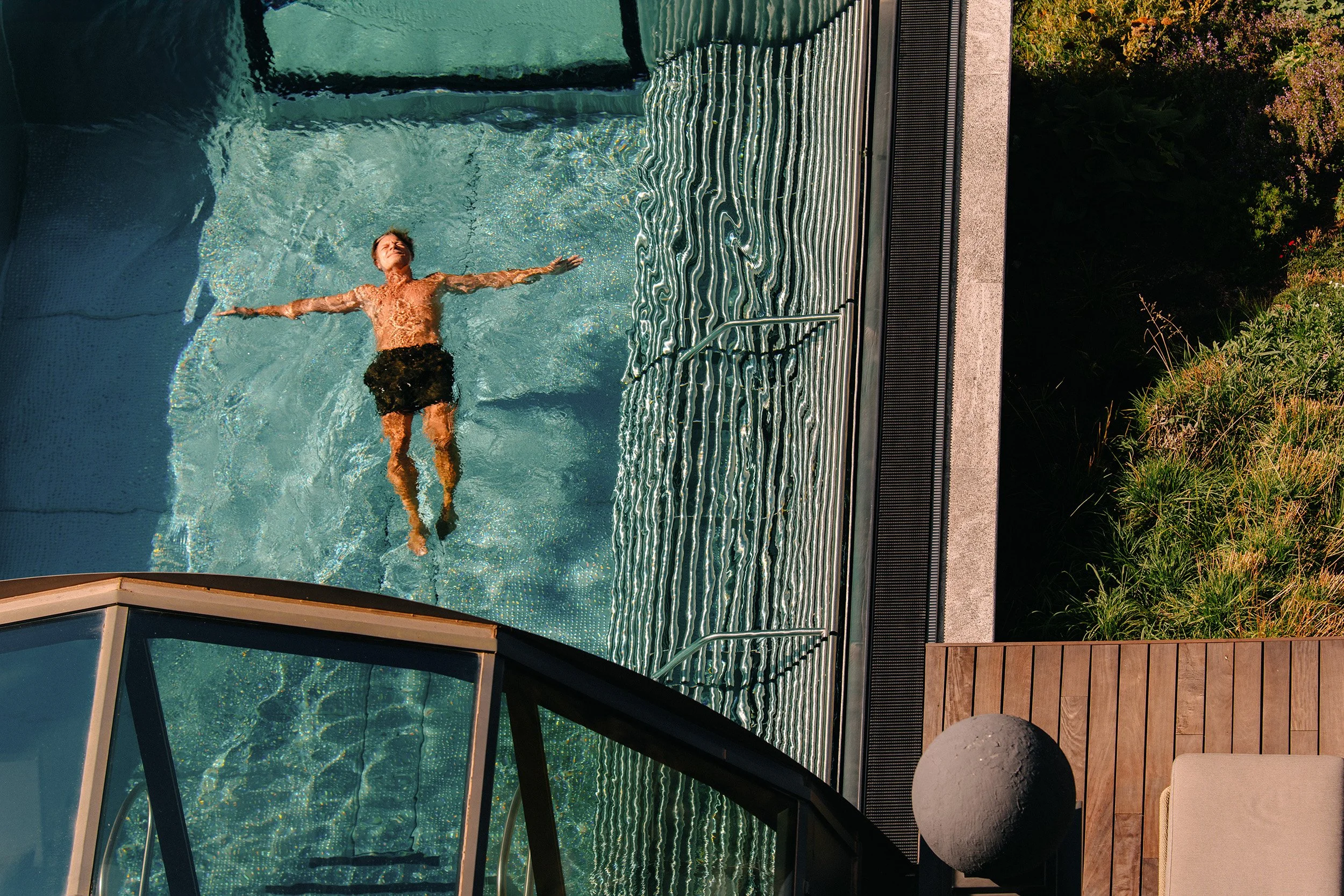 A man floating in a swimming pool with arms outstretched, surrounded by clear water and a metal pool ladder. The pool area has wooden decking and nearby landscaping with plants and bushes.