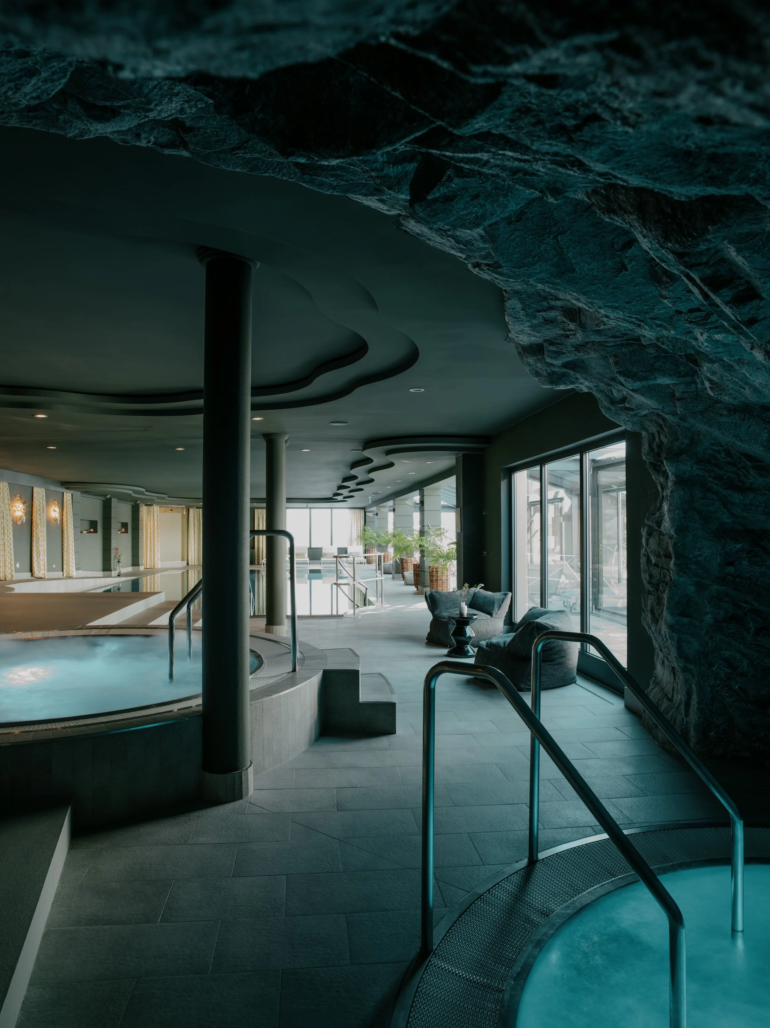 Indoor swim spa area with a circular hot tub, lounge chairs, large glass windows, and artificial rock wall.