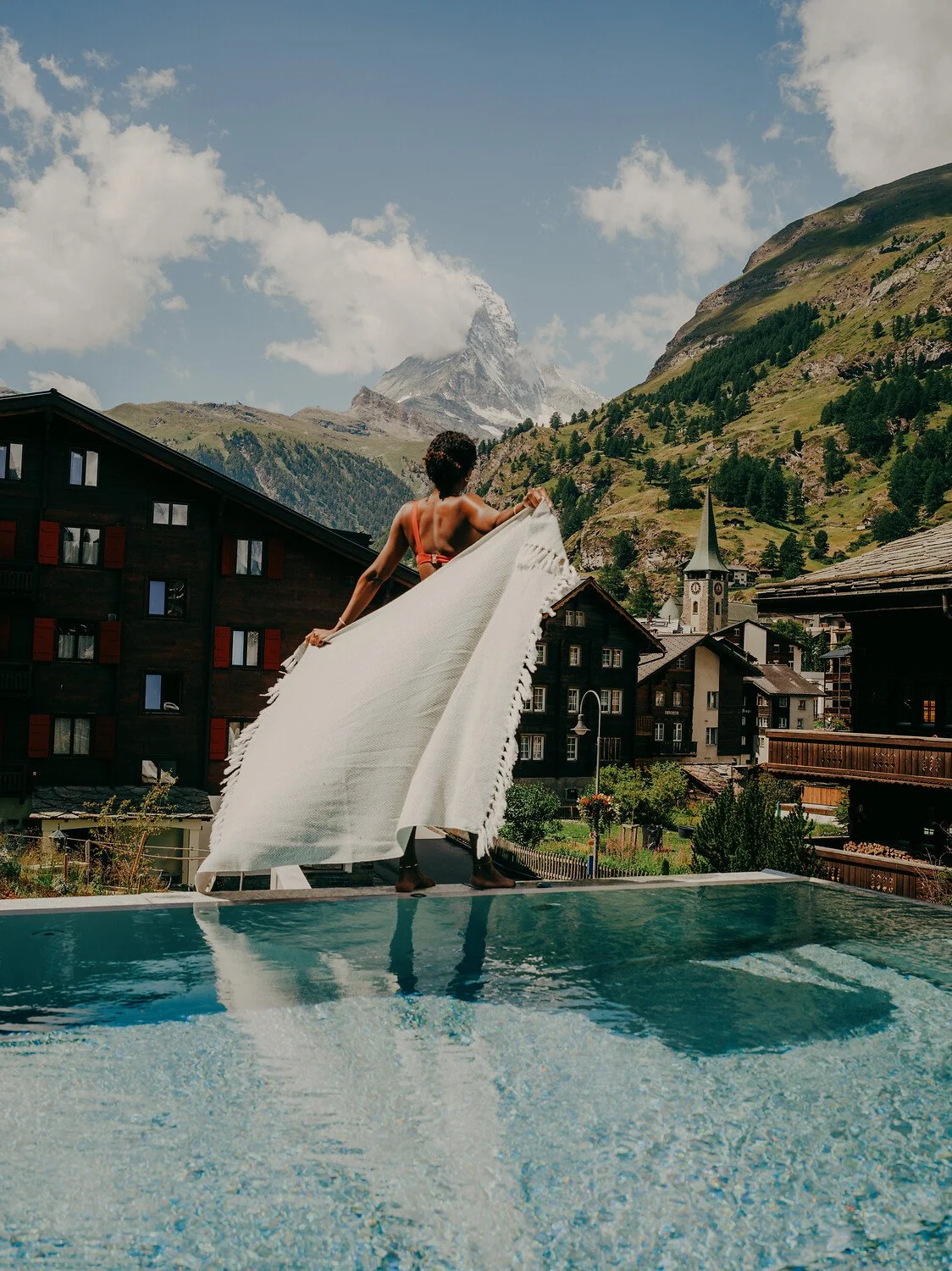 A woman standing at the edge of a swimming pool with a scenic mountain village and snow-capped peaks in the background, holding a white towel or blanket.