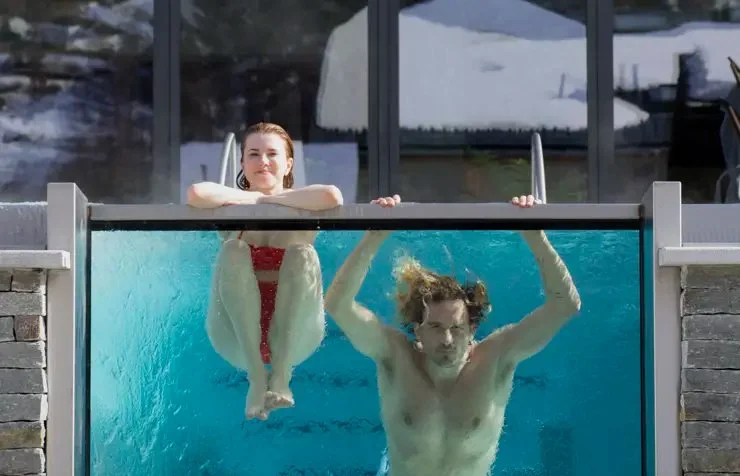 A woman and a man are in a swimming pool, with the woman sitting on the edge above the water and the man hanging upside down in the pool, holding onto the edge.